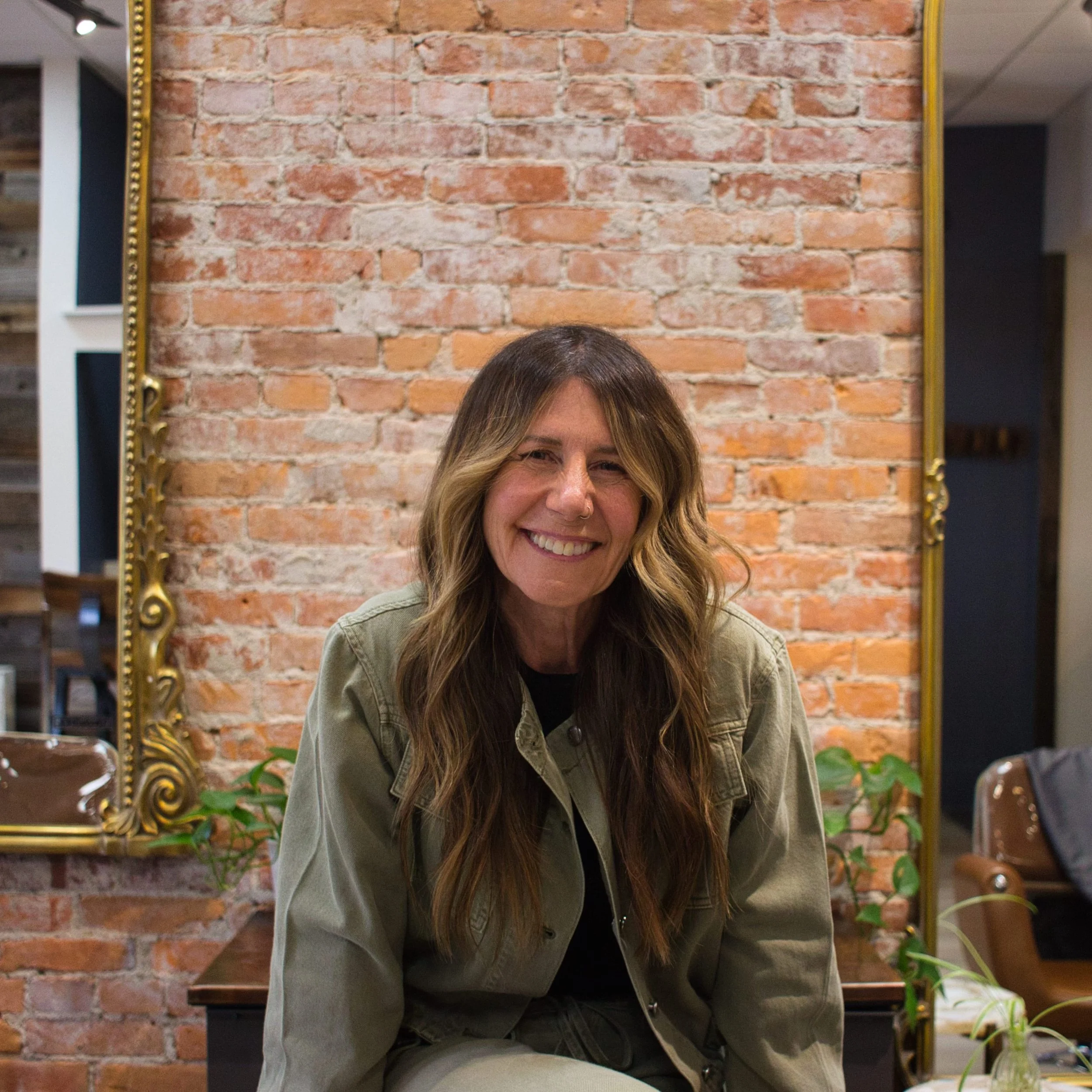 A woman with long, wavy hair smiling, sitting in front of an exposed brick wall with a gold-framed mirror on her left and potted plants around her.