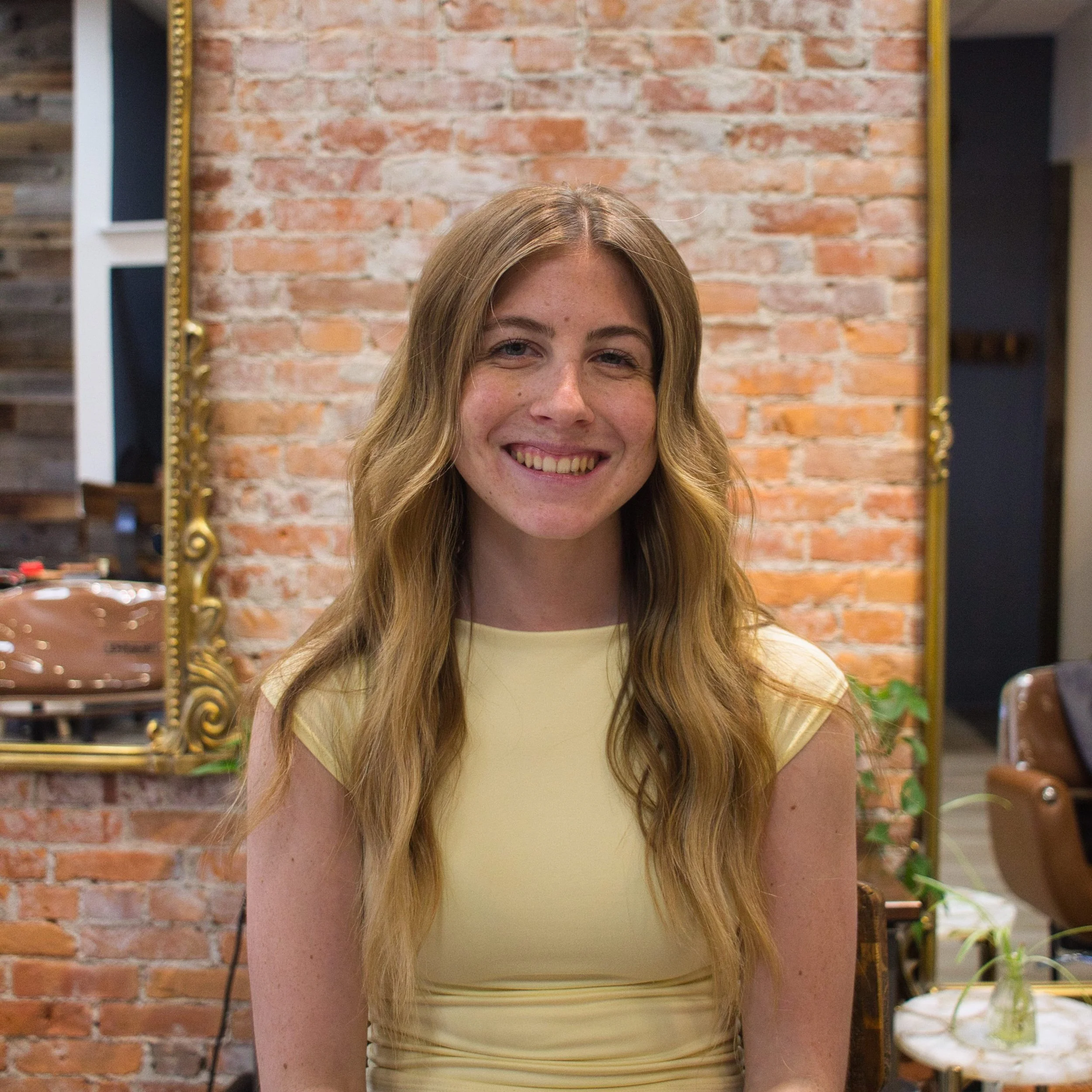 A young woman with long blonde hair smiling in front of a brick wall, with a gold-framed mirror and shelves with wooden boxes and plants in the background.