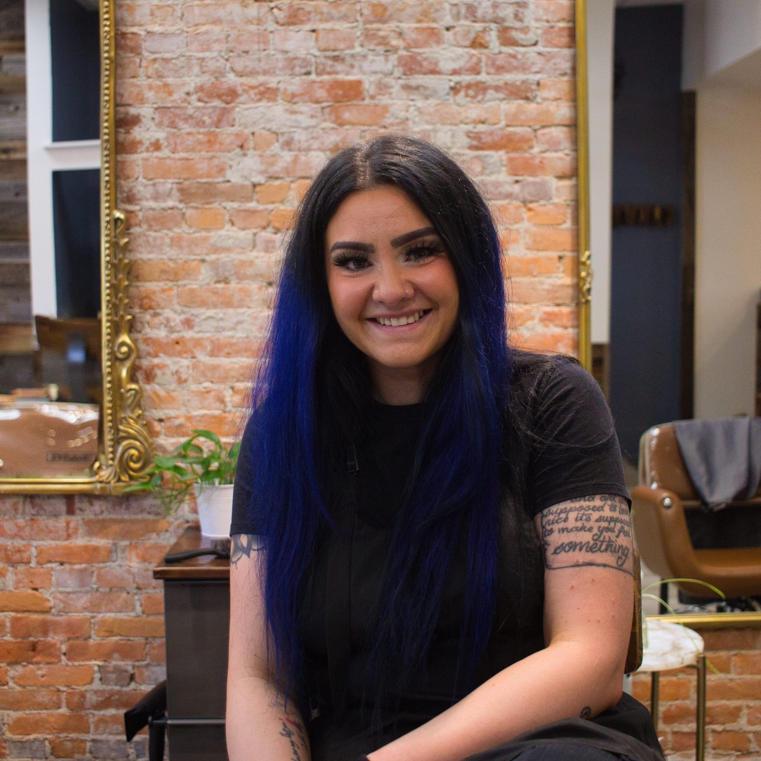 A young woman with long black hair, blue tips, and tattoos on her arm, smiling in front of a brick wall and a mirror with a gold frame.