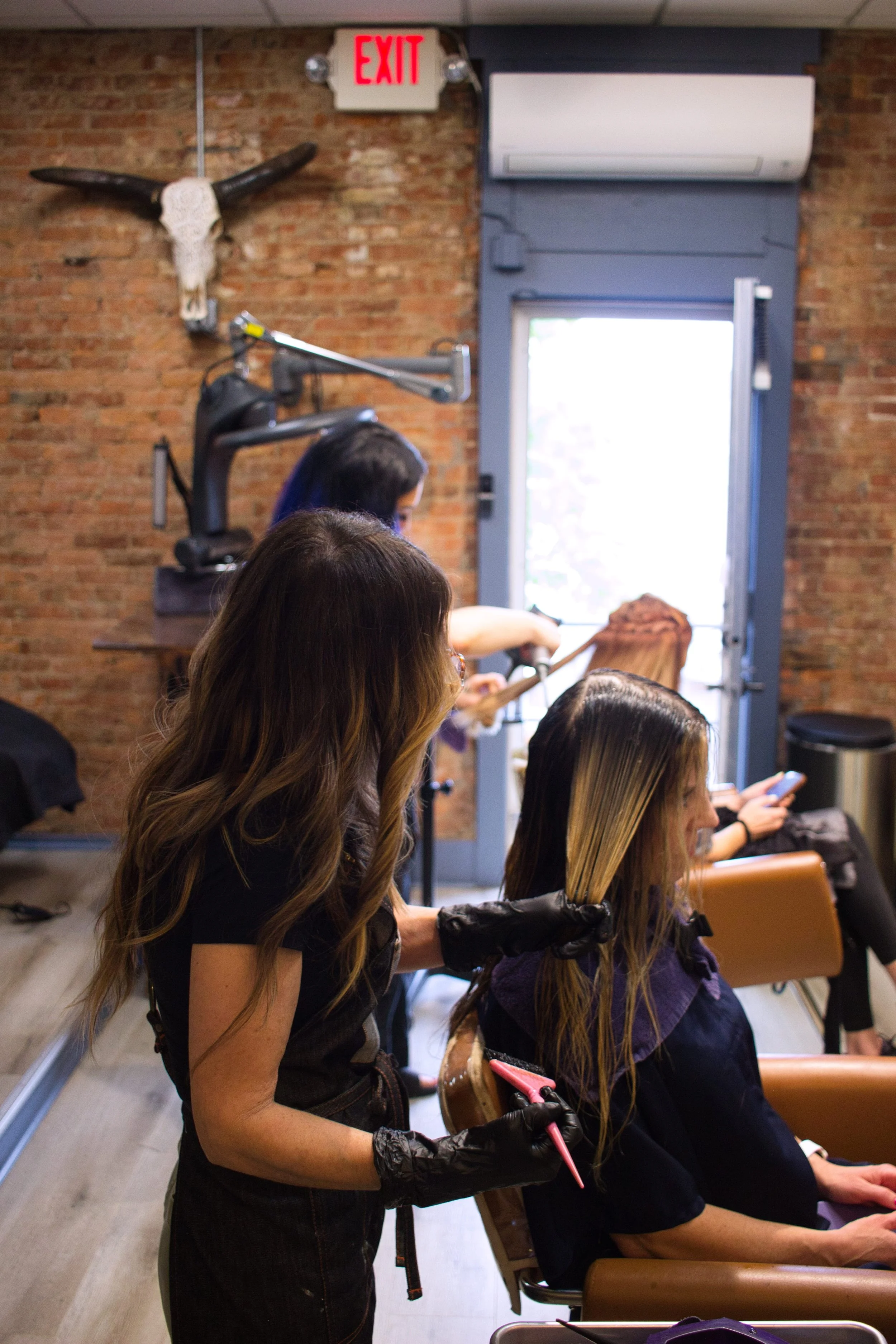 A young woman is using pink scissors to cut a woman's long, blonde hair in a salon, while other customers wait seated with purple hair coverings and black gloves. The salon has a brick wall, an aircraft cow skull mounted, and an open door with sunlight outside.