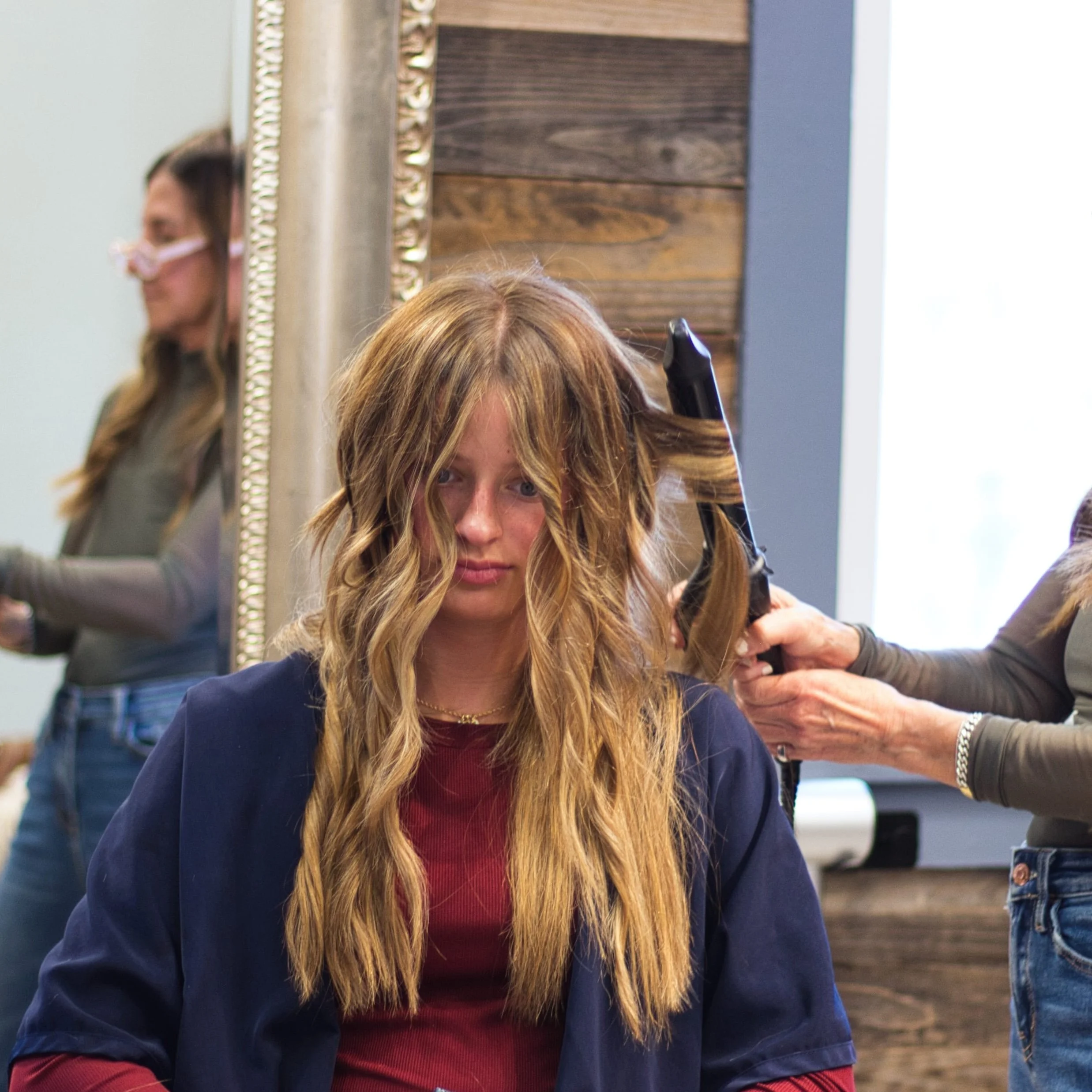 A woman with long blonde hair, wearing a maroon top and dark jacket, is in a hair salon getting her hair styled or cut by a stylist. The stylist's arm and hand are visible, holding a curling iron. There is a mirror with decorative border behind her, and another person with long dark hair and a face mask is reflected in the mirror.