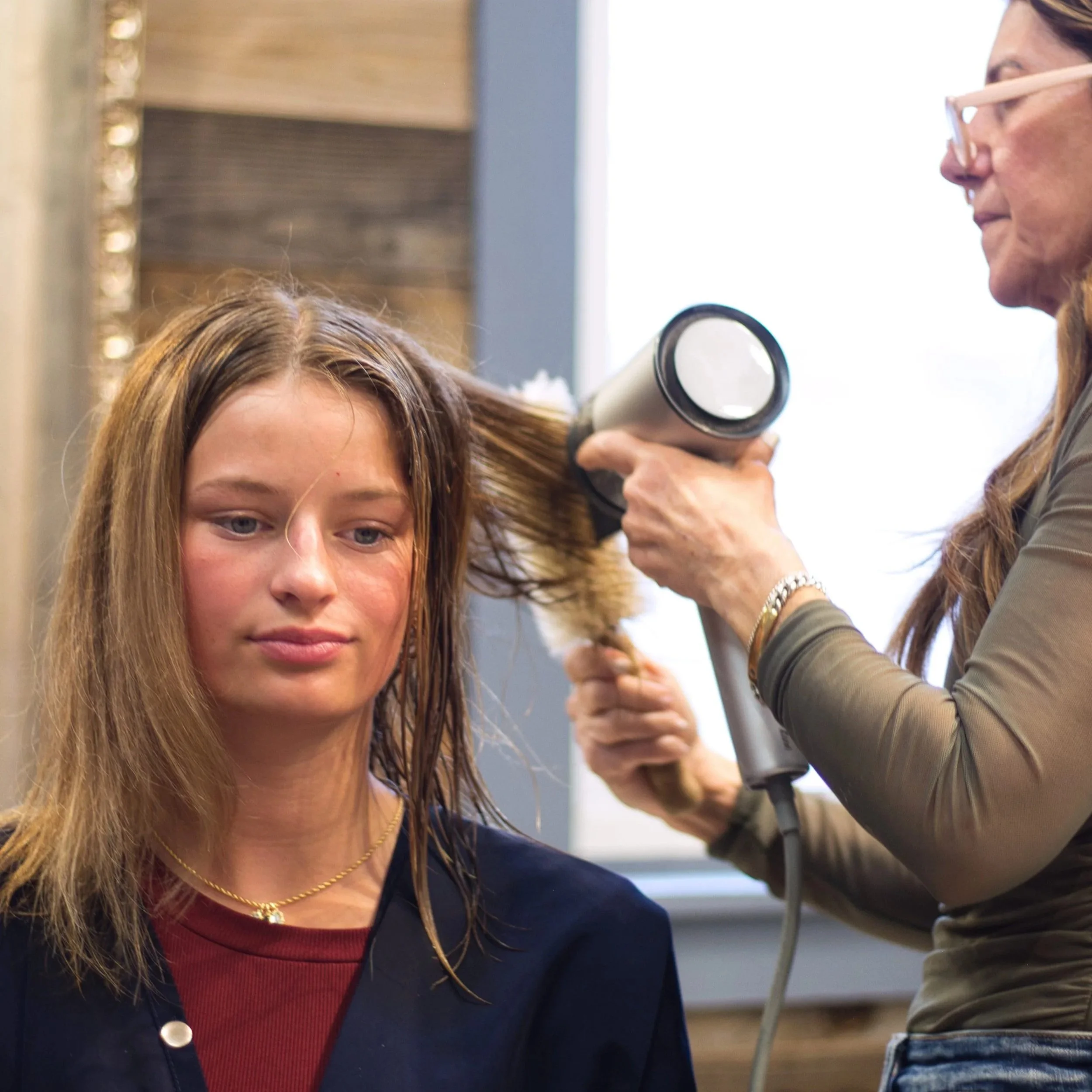 A woman with light brown, wavy hair using a hairdryer on a younger woman with straight, light brown hair in a hair salon.