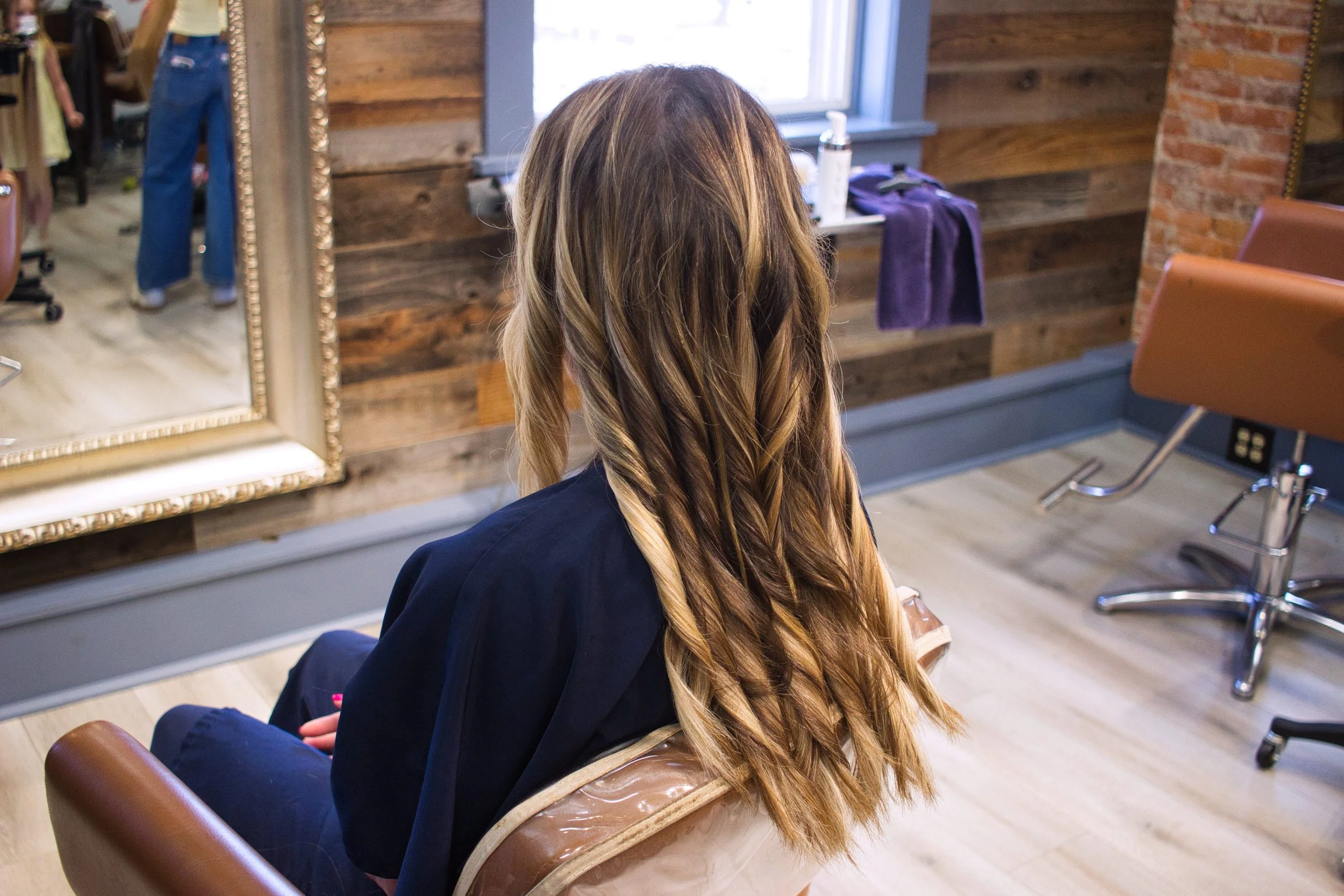 Back of a woman with long wavy blonde hair sitting in a hair salon chair, facing a mirror, with a wooden wall and window in the background.