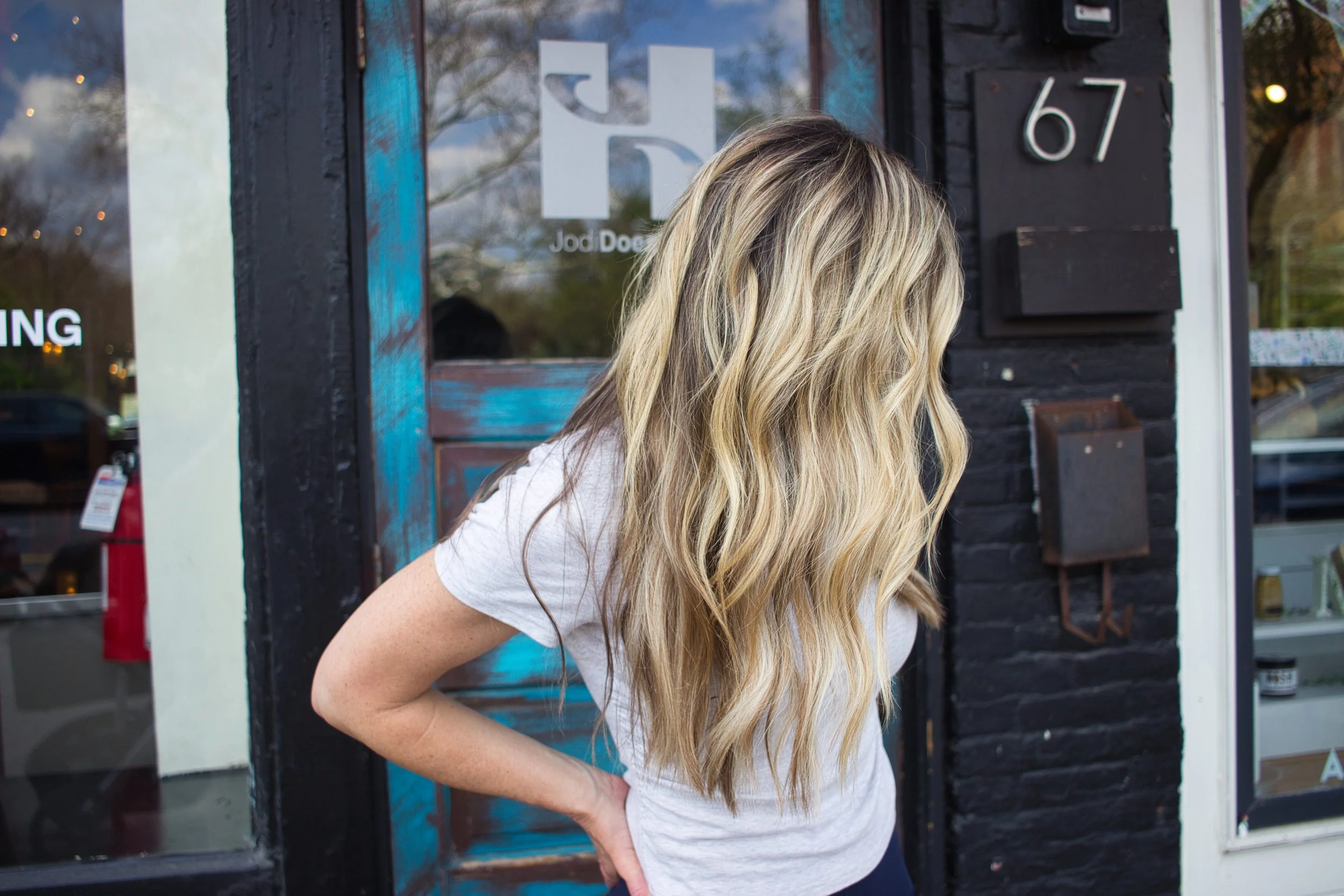 A woman with long, wavy blonde hair wearing a light gray t-shirt standing outside a building with the number 67 on a black brick wall.
