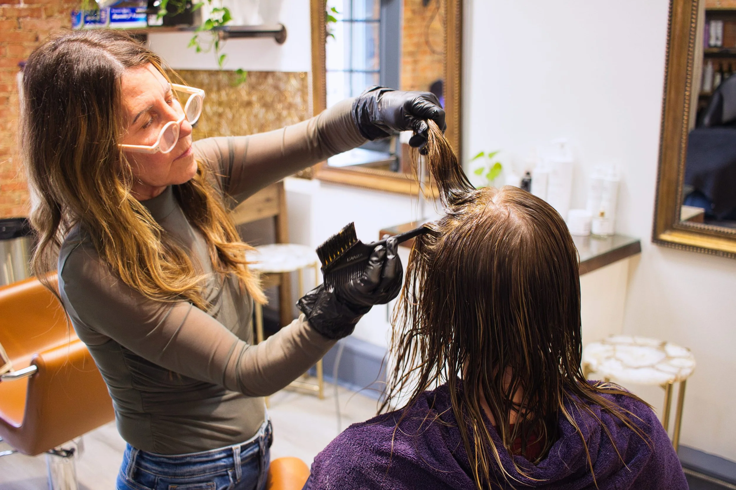 A hairstylist with long hair, glasses, and black gloves is applying hair dye or treatment to a woman's wet hair in a salon. The woman is seated with a purple towel around her shoulders, and the salon features a large mirror, exposed brick wall, and various hair products.