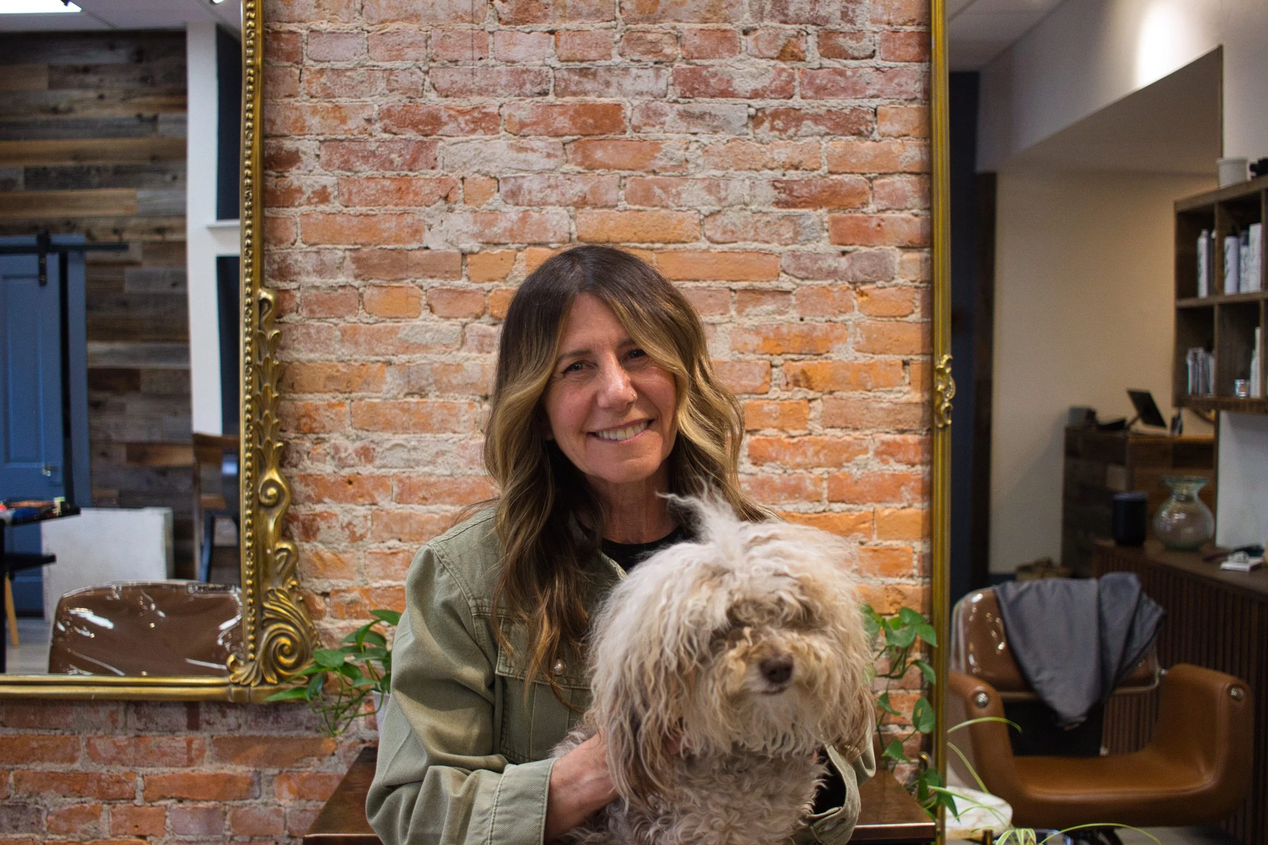 A woman with wavy brown hair smiling while holding a light-colored fluffy dog in front of a brick wall and a large mirror.