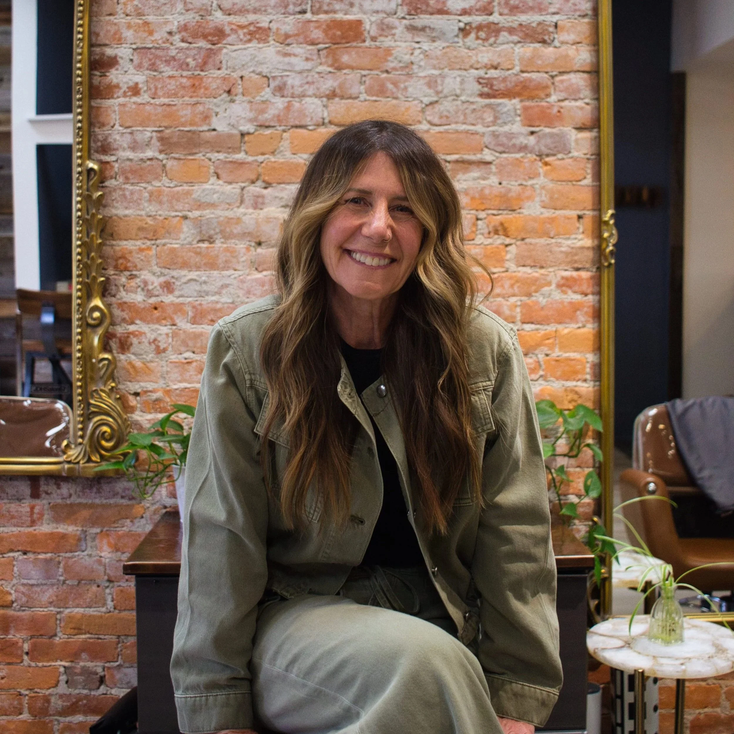 A woman with long wavy hair smiling, sitting in front of a brick wall with a large mirror frame to her side, and a small table with plants and a drink nearby.