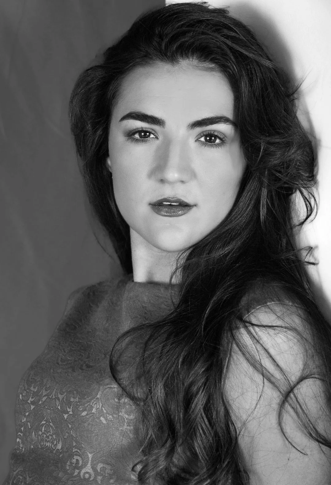 Black and white portrait of a woman with long, wavy hair, wearing a lace top, looking directly at the camera.