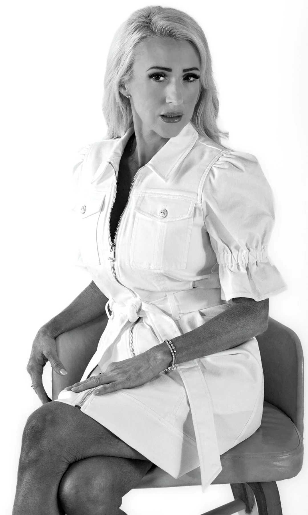 Black and white photo of a woman with blonde hair sitting on a chair against a plain white background. She is wearing a light-colored dress with puffed sleeves and a tie at the waist, and she has jewelry on her wrist and ears. She has a confident exp