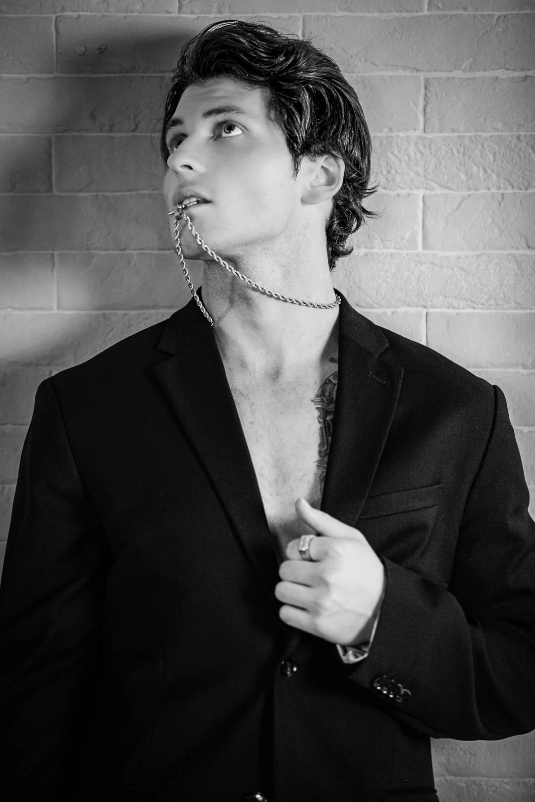 A young man in a black suit jacket, no shirt underneath, with a chain in his mouth and tattoos visible on his chest. He is posing against a brick wall, looking upwards, with one hand adjusting his jacket.