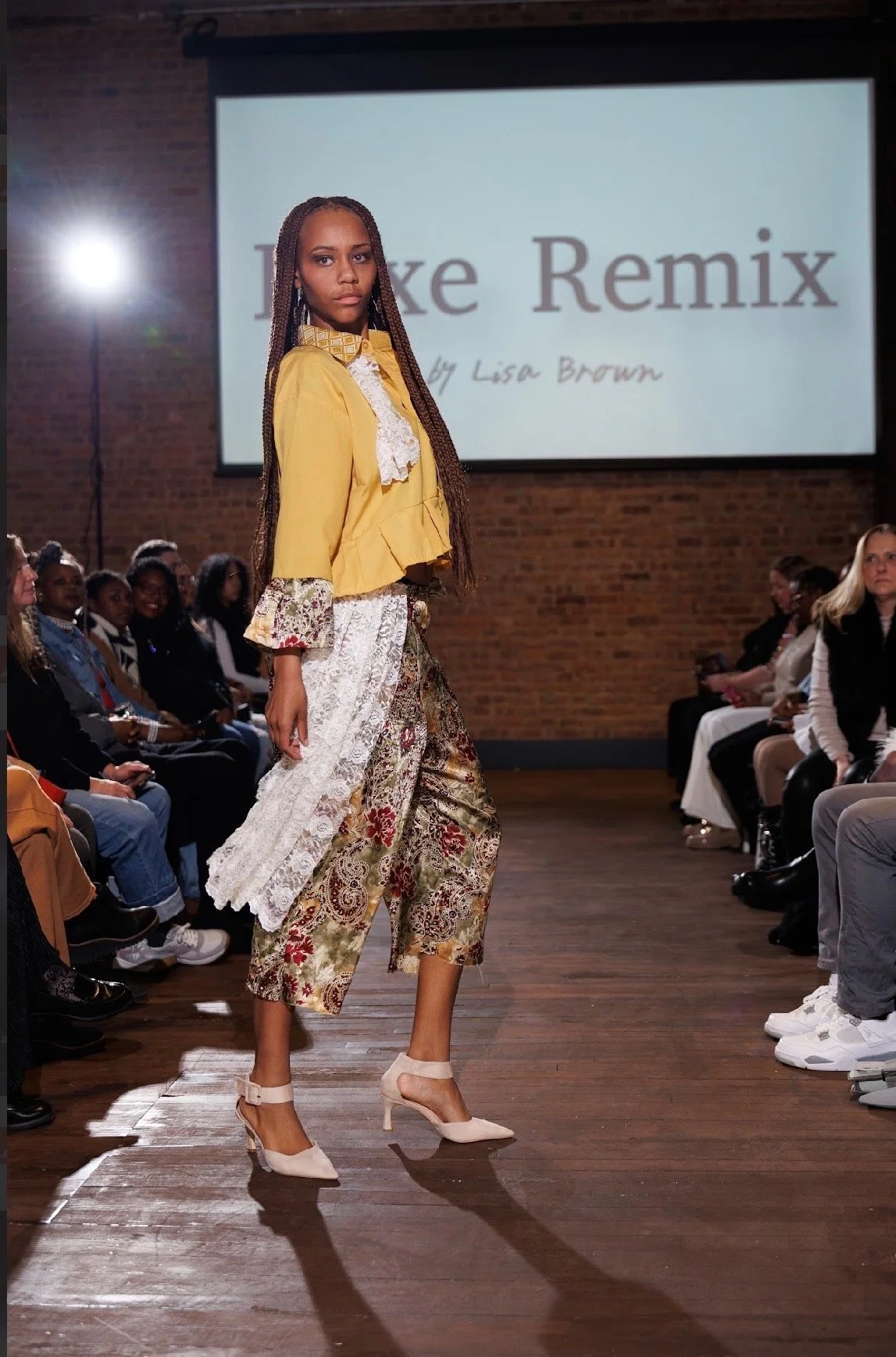 A model walks down the runway at a fashion show, wearing a yellow jacket with lace details and floral cropped pants with lace accents. The audience is seated on either side of the runway, and a large screen in the background displays the text 'Luxe R