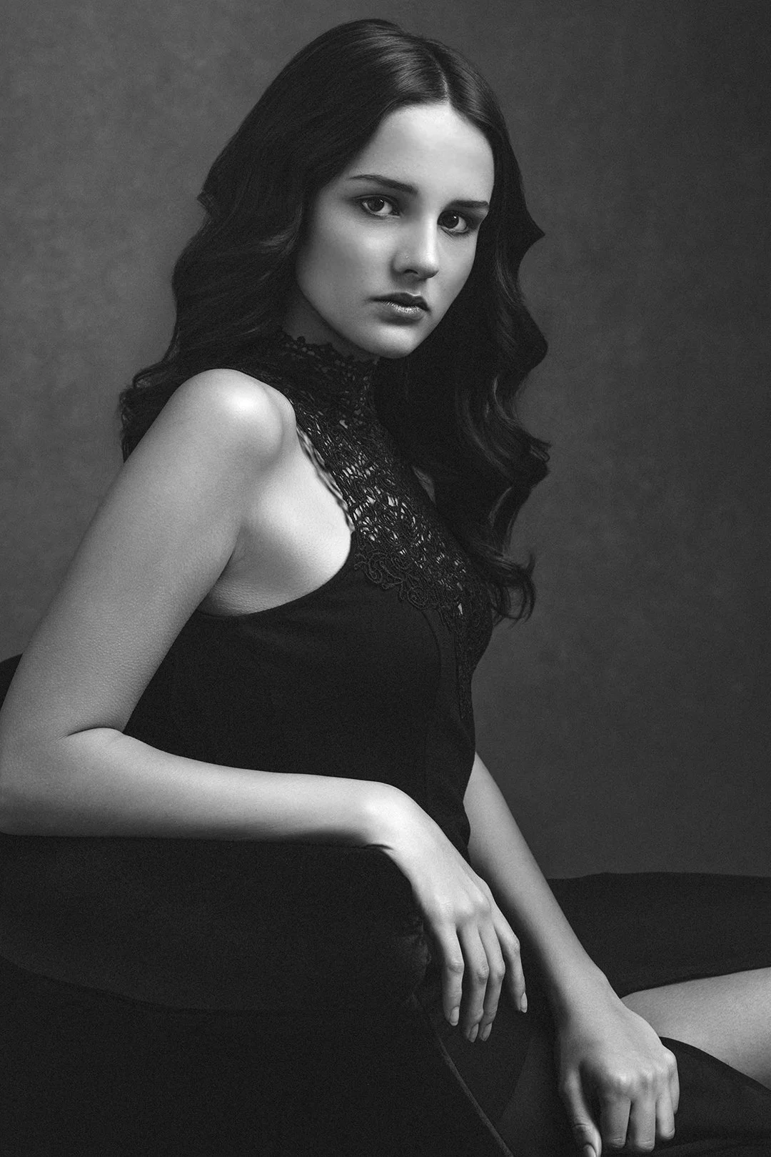 Black and white portrait of a young woman with long dark wavy hair wearing a sleeveless lace top, sitting against a plain dark background.