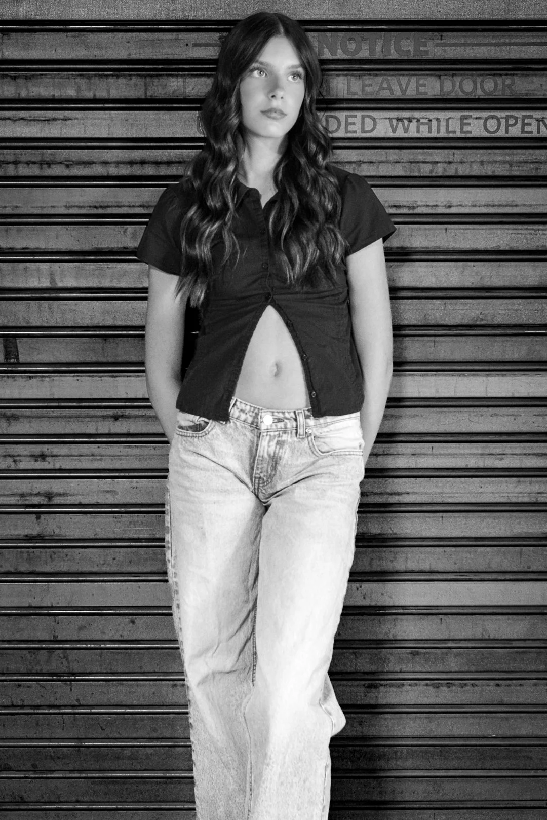 A woman with long wavy hair stands against a metal garage door, partially lifting her unbuttoned black shirt to reveal her midriff, wearing light-wash jeans.