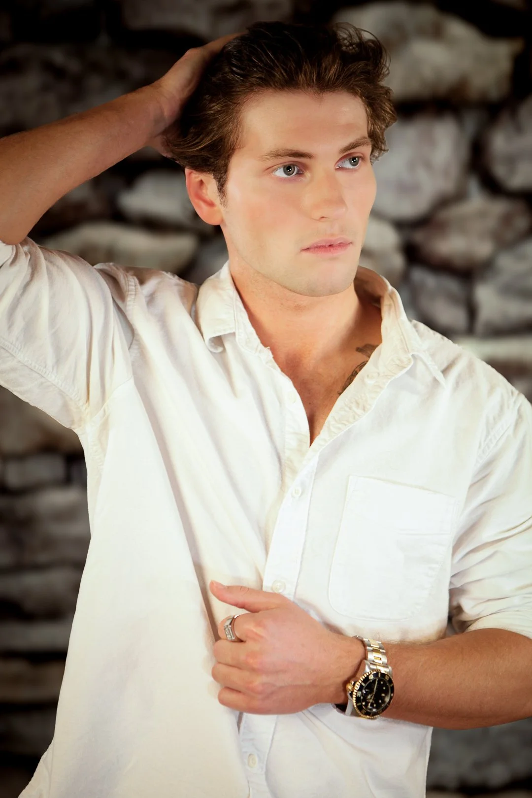 A young man with light skin, blue eyes, and brown hair is wearing a white button-up shirt and a large wristwatch. He has his left hand on his head and is looking into the distance against a background of rocks.