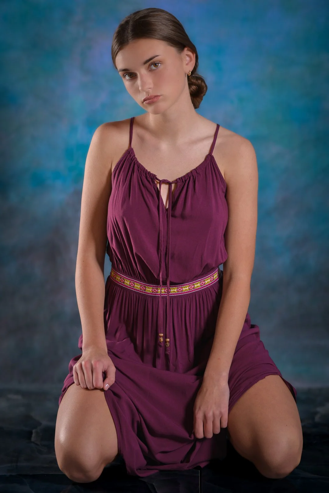A young woman with brown hair tied in a low bun, sitting on her knees against a blue textured background. She is wearing a purple sleeveless dress with thin straps and decorative stitching at the waist, looking slightly to her left with a neutral exp
