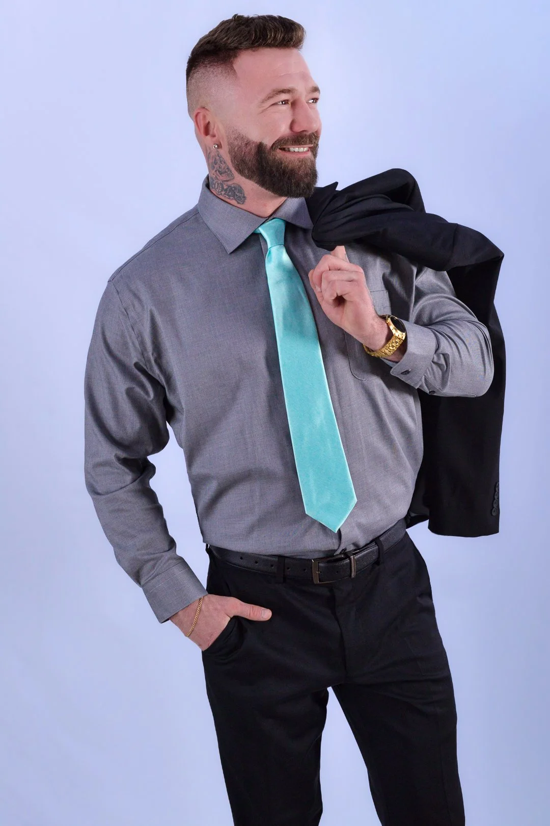 A man in a gray dress shirt and black pants holding a black blazer over his shoulder, smiling, with a turquoise tie and gold watch, posing against a light background.