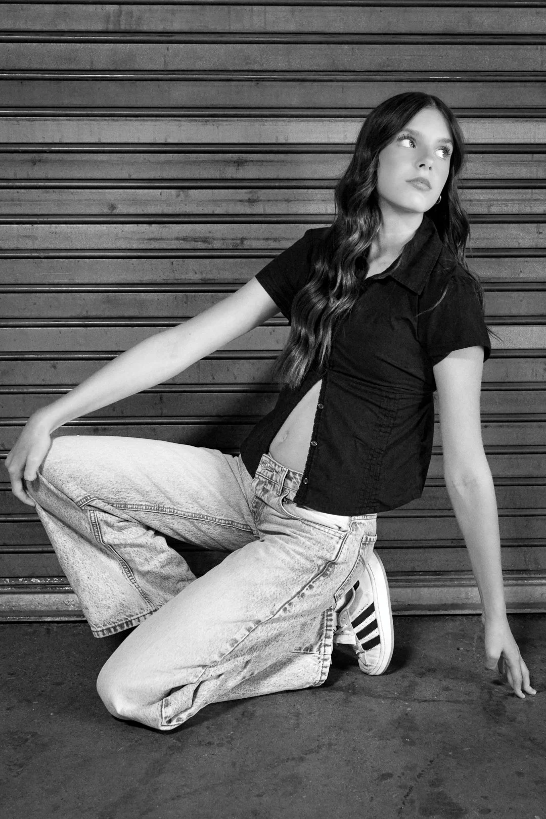 Black and white photo of a woman in casual attire, sitting on the ground with a wooden wall behind her, looking to the side.
