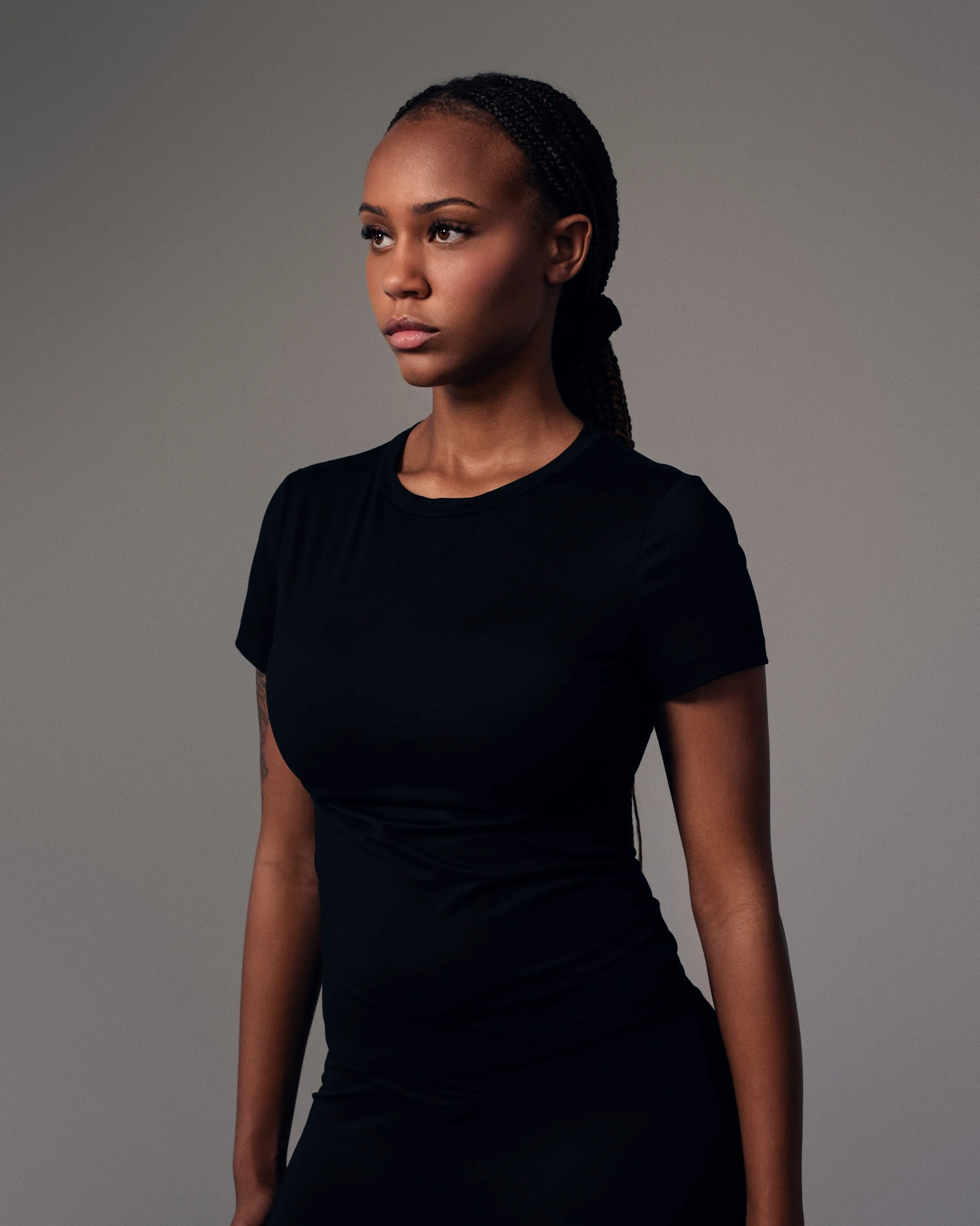 Portrait of a young Black woman with braided hair wearing a black shirt against a plain gray background.