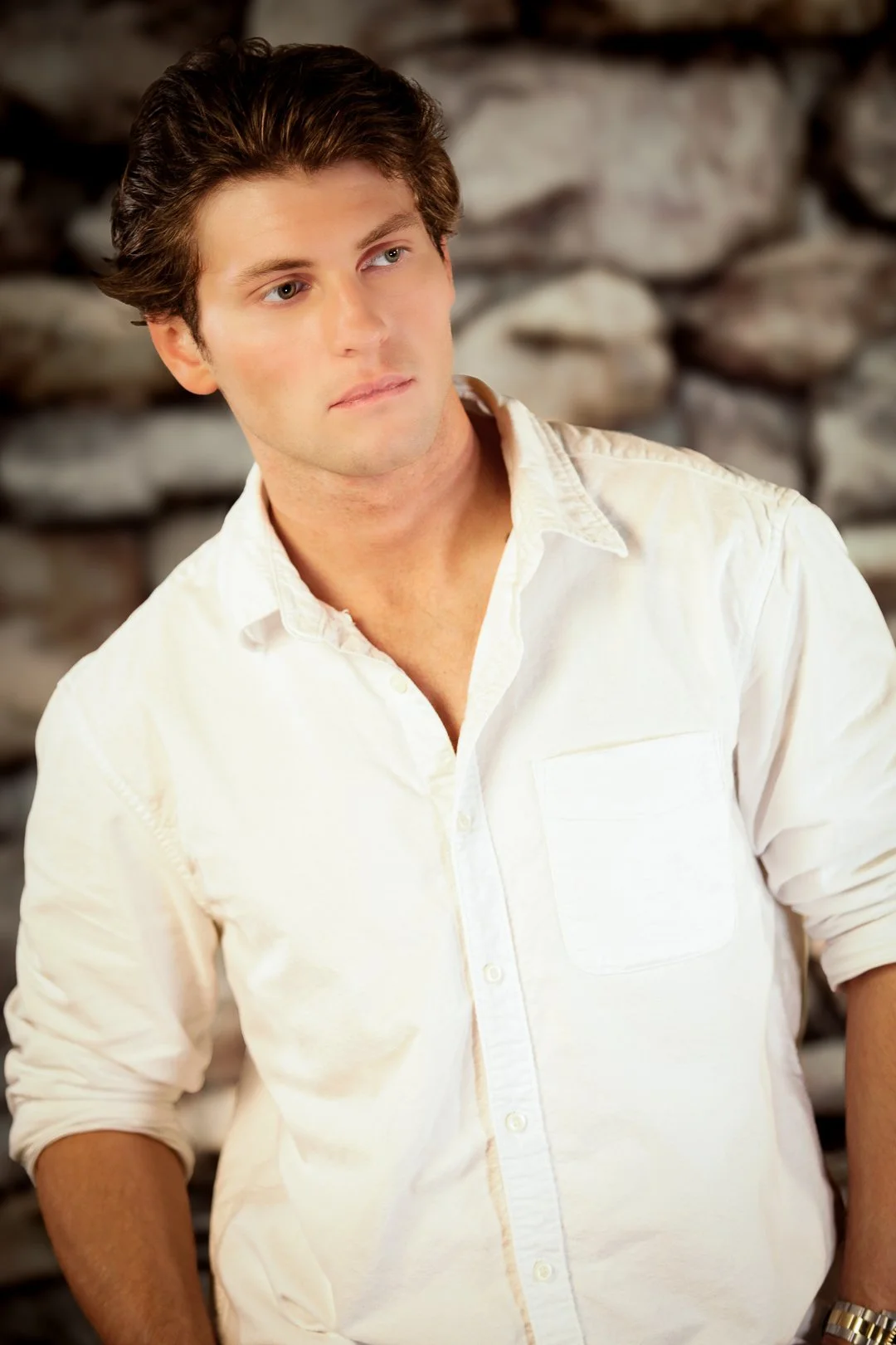 A young man with brown hair, wearing a white button-up shirt, standing in front of a stone wall.