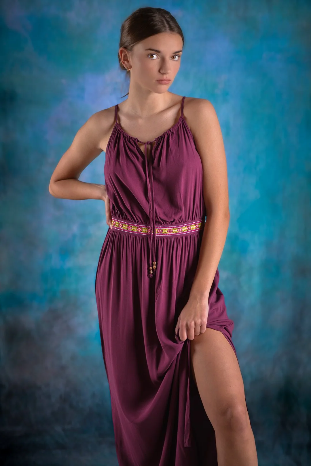 A woman with brown hair in a purple dress with thin straps and an embroidered waistband, standing against a colorful, textured blue background, lifting one side of her dress to reveal her thigh.