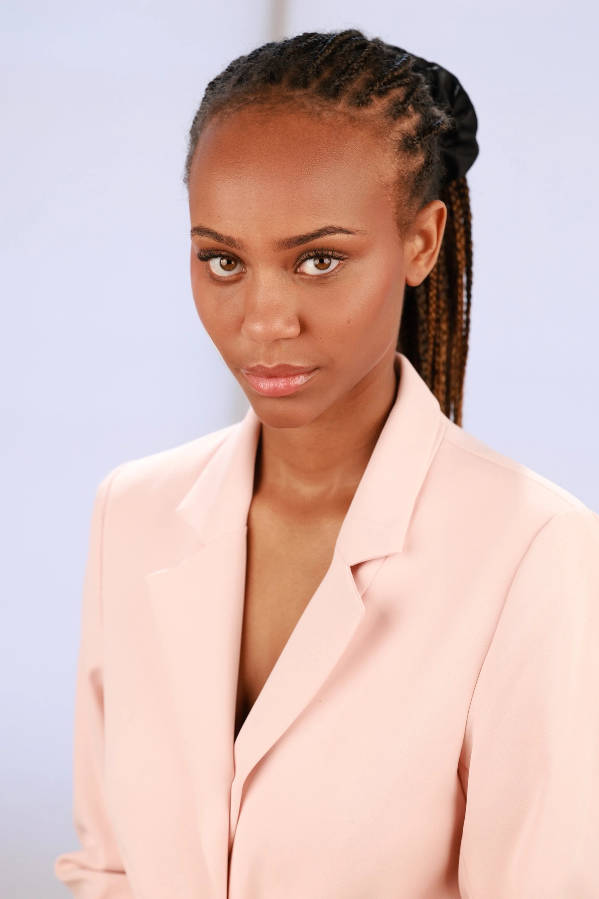 A woman with dark skin and blonde braids wearing a light pink blazer against a plain light background.