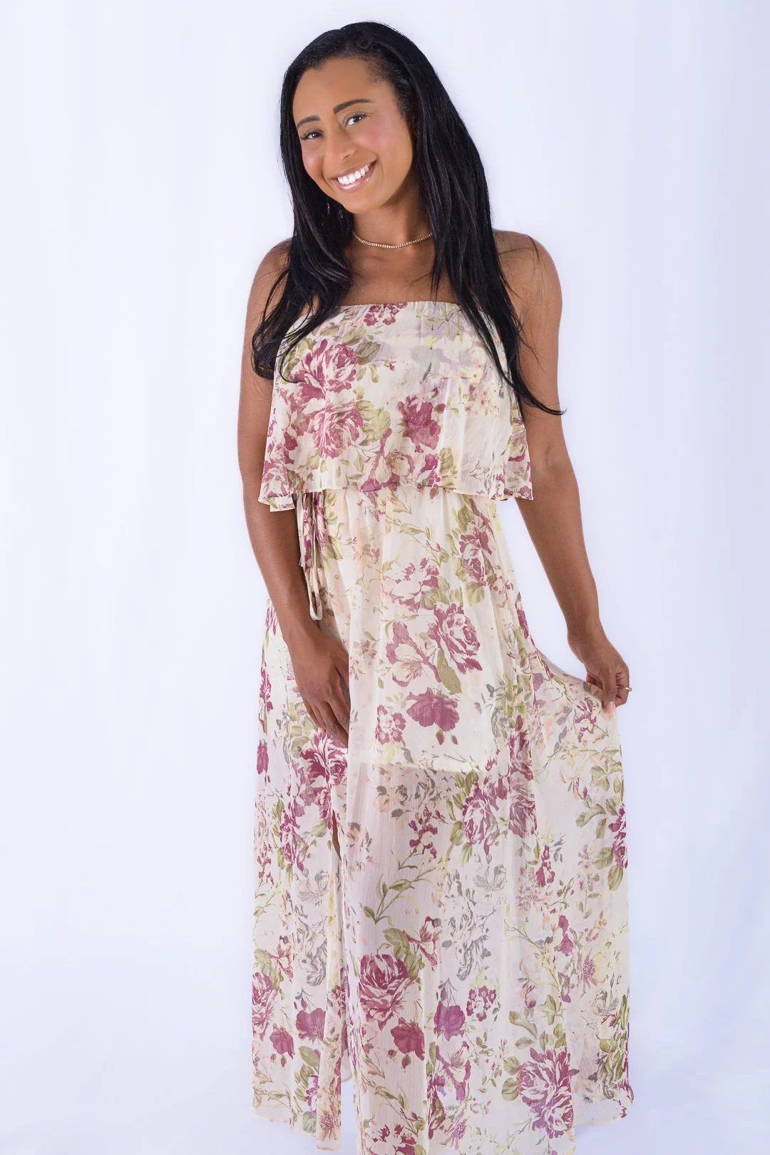 A young woman with long black hair wearing a sleeveless, floral print maxi dress in shades of pink, green, and cream, smiling and standing against a plain white background.
