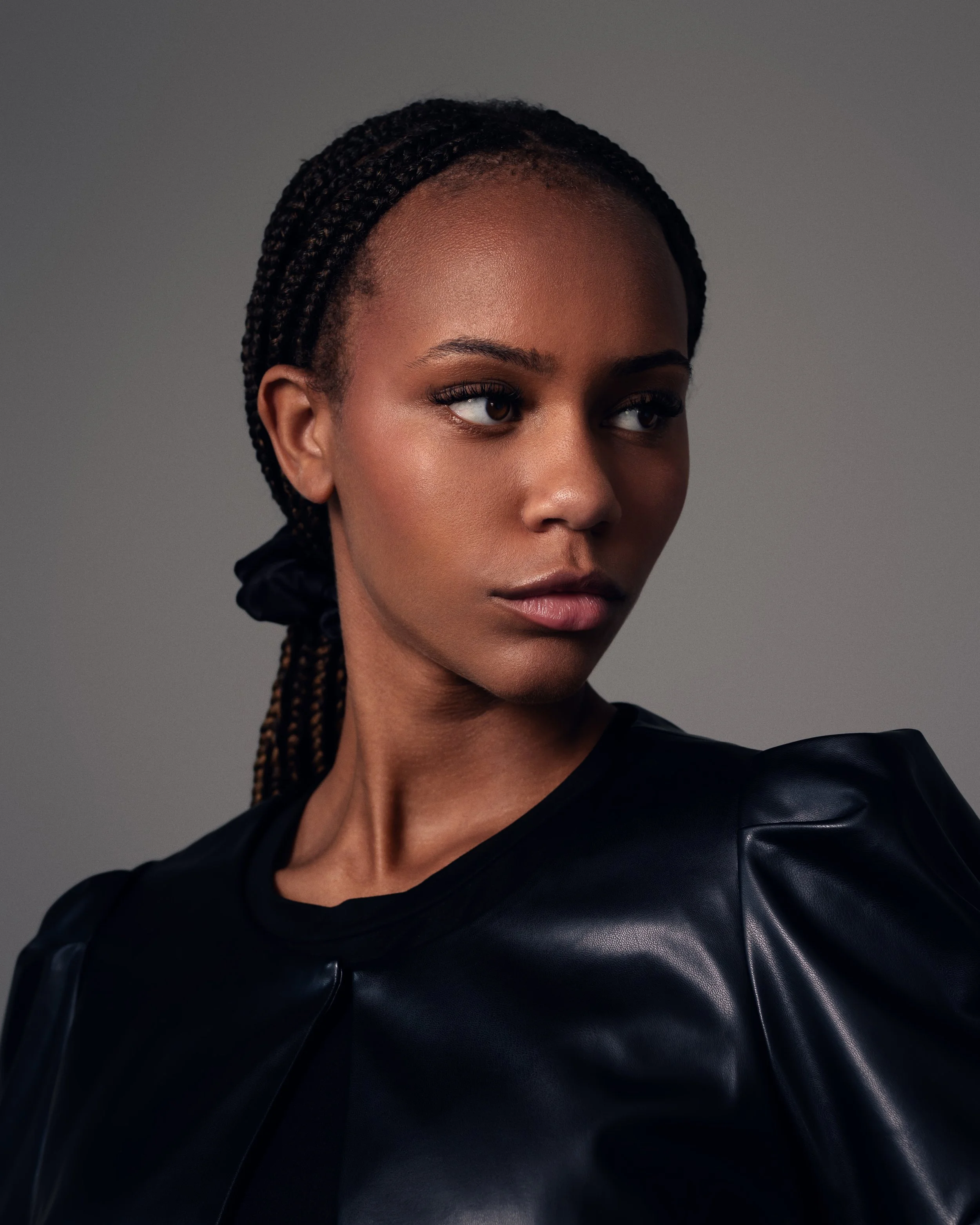 A woman with braids wearing a black leather jacket and black top, looking to her right against a gray background.