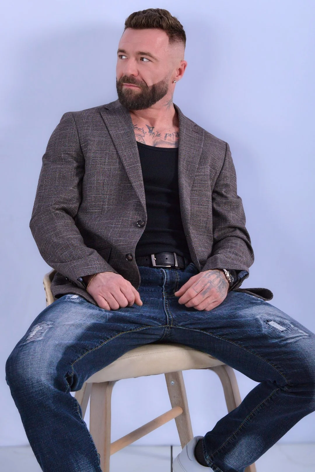 A man with a beard, tattoos, and earrings sitting on a wooden stool against a light background, wearing a blazer, black shirt, distressed jeans, and white sneakers.