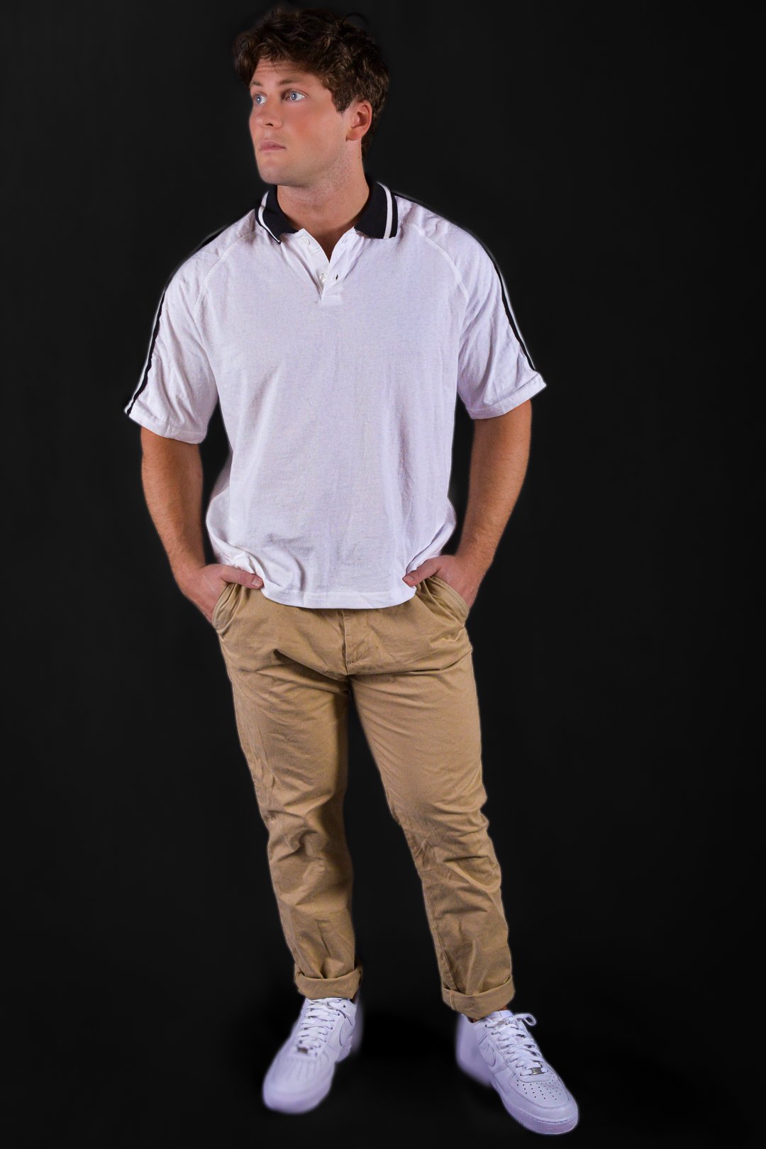 A young man with light skin and brown hair standing against a black background, wearing a white polo shirt with black trim, tan pants, and white sneakers.