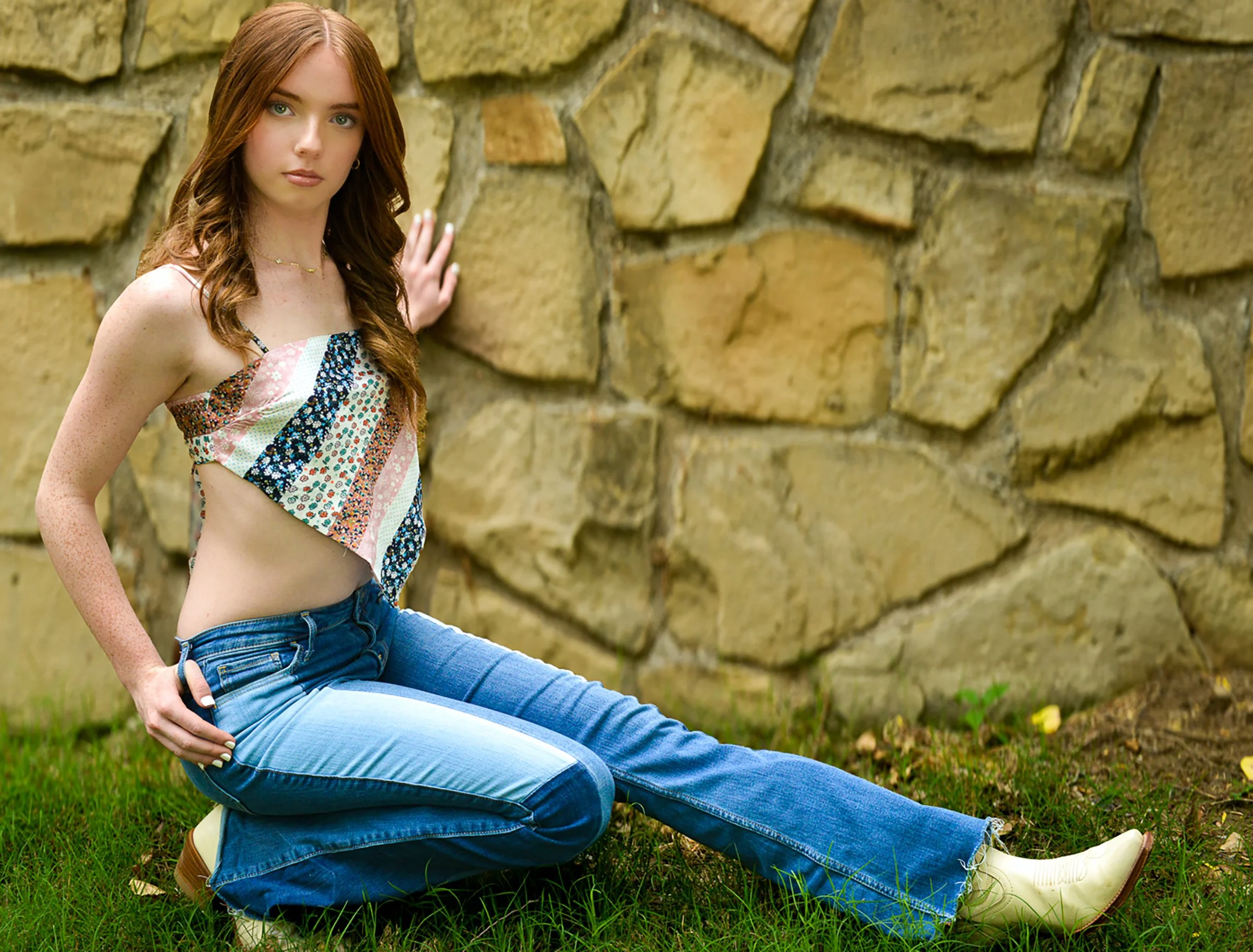 A woman with red hair in a colorful tank top and blue jeans sitting on the grass beside a stone wall, looking at the camera.