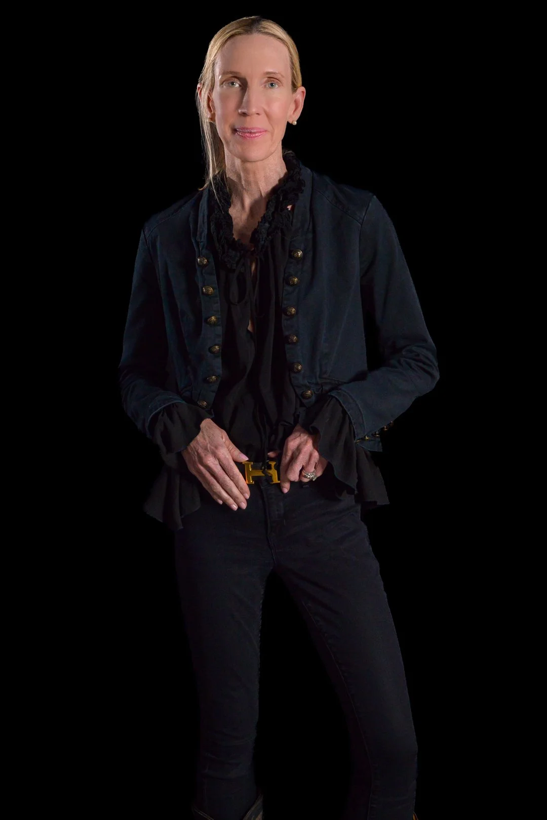 A woman with long blonde hair wearing a black jacket, black pants, and a black blouse with ruffled trim, standing against a black background.