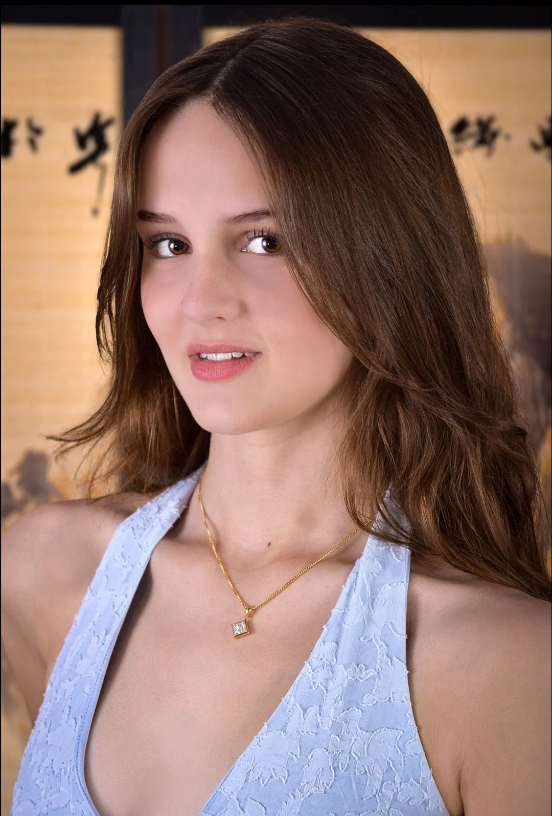 A young woman with light skin and long, wavy brown hair, wearing a blue lace top and a gold necklace with a square-shaped pendant, standing in front of a traditional Asian screen with Chinese characters.