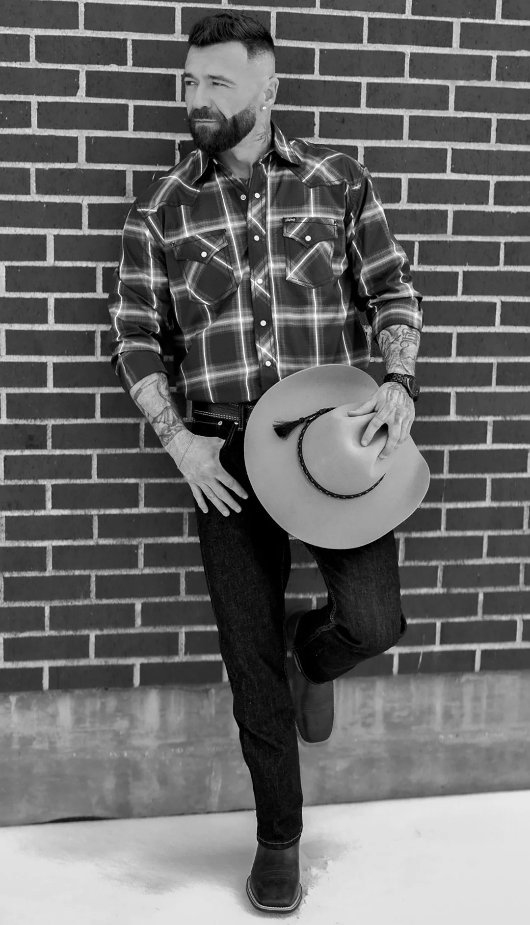 A man with a beard and tattoos wearing a plaid shirt, jeans, and cowboy boots holding a wide-brimmed hat with a rope hatband, standing against a brick wall.