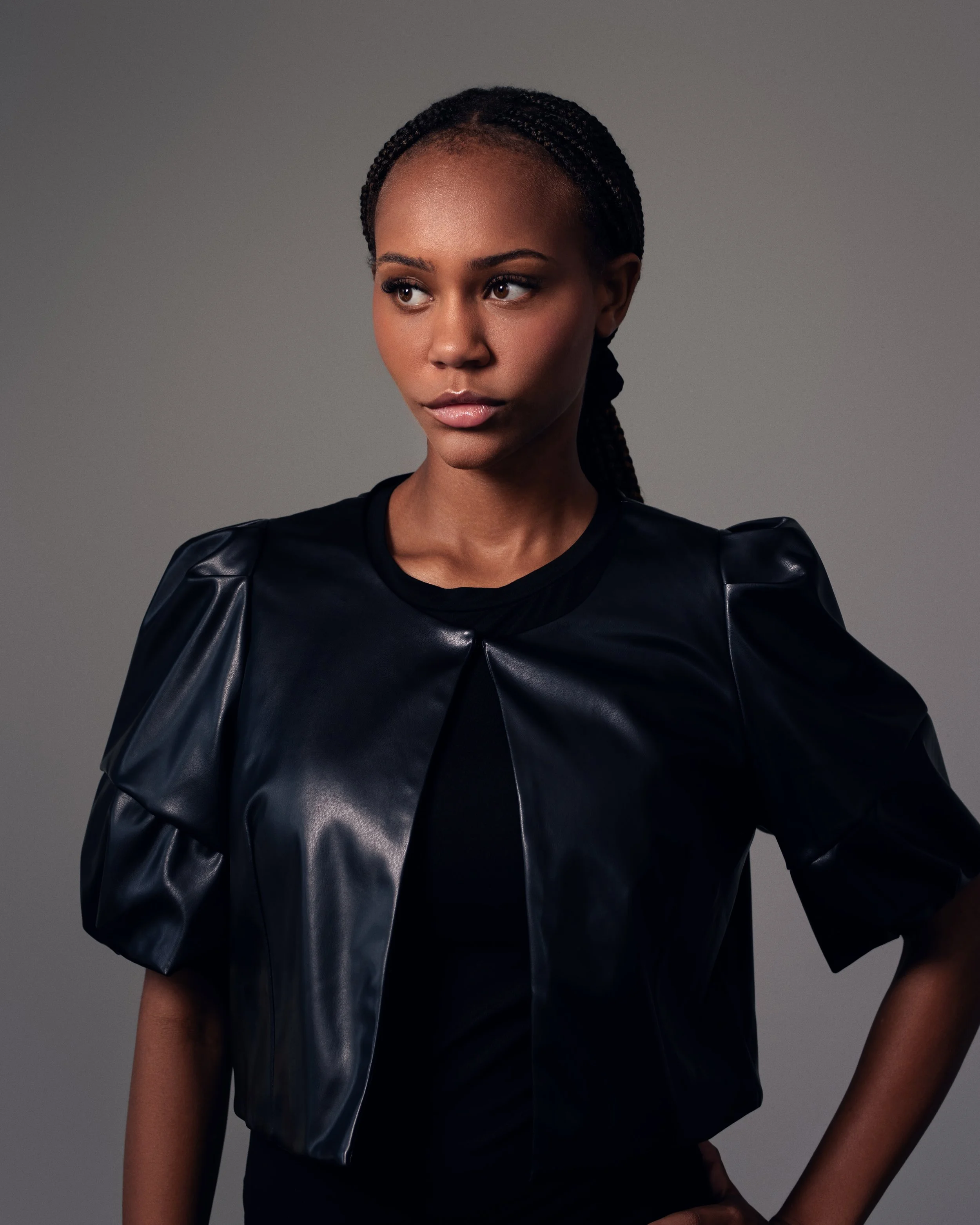 A woman with braided hair wearing a black leather jacket against a gray background.