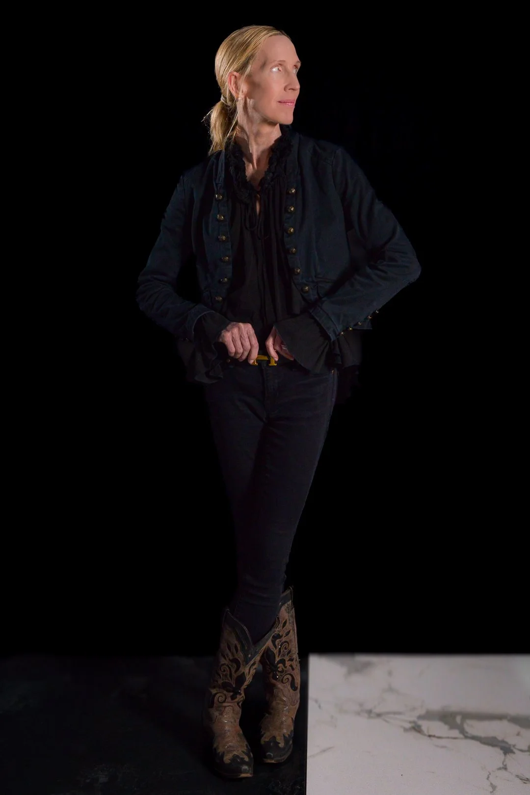 A woman with blonde hair tied in a ponytail, wearing a dark jacket, black shirt with ruffles, black pants, and cowboy boots, standing against a black background and looking to her right.