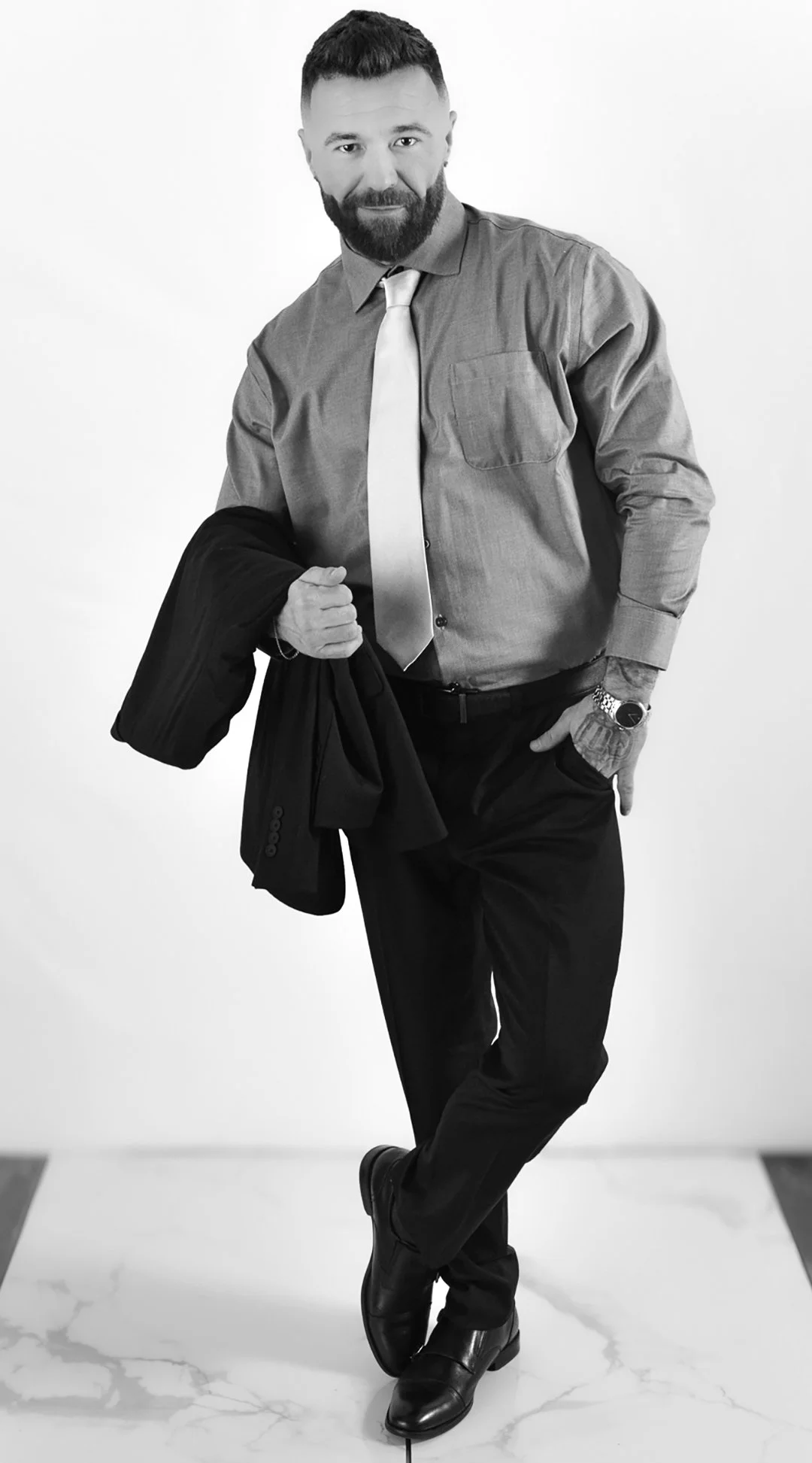 A man with a beard and short hair, dressed in a collared shirt, tie, and black pants, holding a jacket in his right hand, standing against a white background.