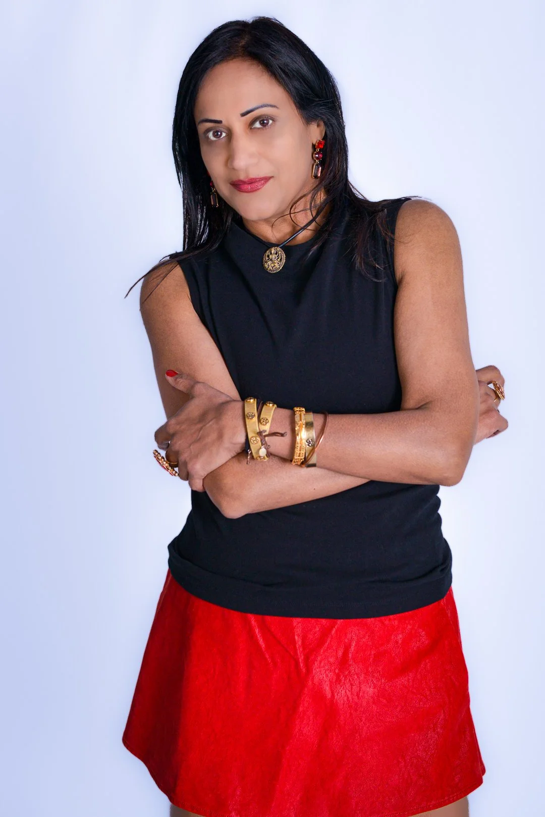 A woman with black hair, wearing a black sleeveless top and red skirt, is standing with arms crossed against a plain white background. She has red and gold jewelry, including earrings, bracelets, and rings.
