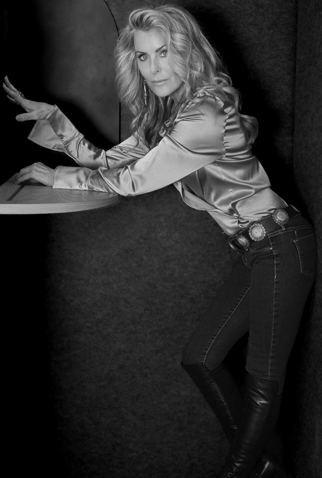 Black and white photo of a woman with wavy blonde hair, wearing a shiny satin blouse with ruffled sleeves, dark jeans, and a decorative belt, leaning forward with her arms extended on a small table or shelf, in a cozy, dark interior space.