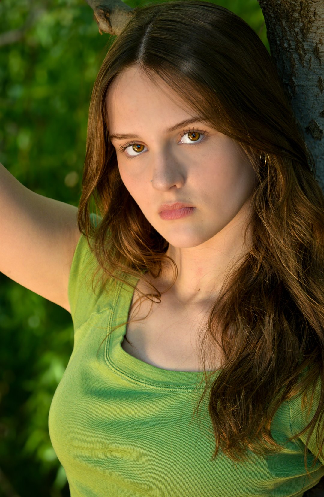 A young woman with brown hair and light skin leaning against a tree outdoors, wearing a green top and looking at the camera.