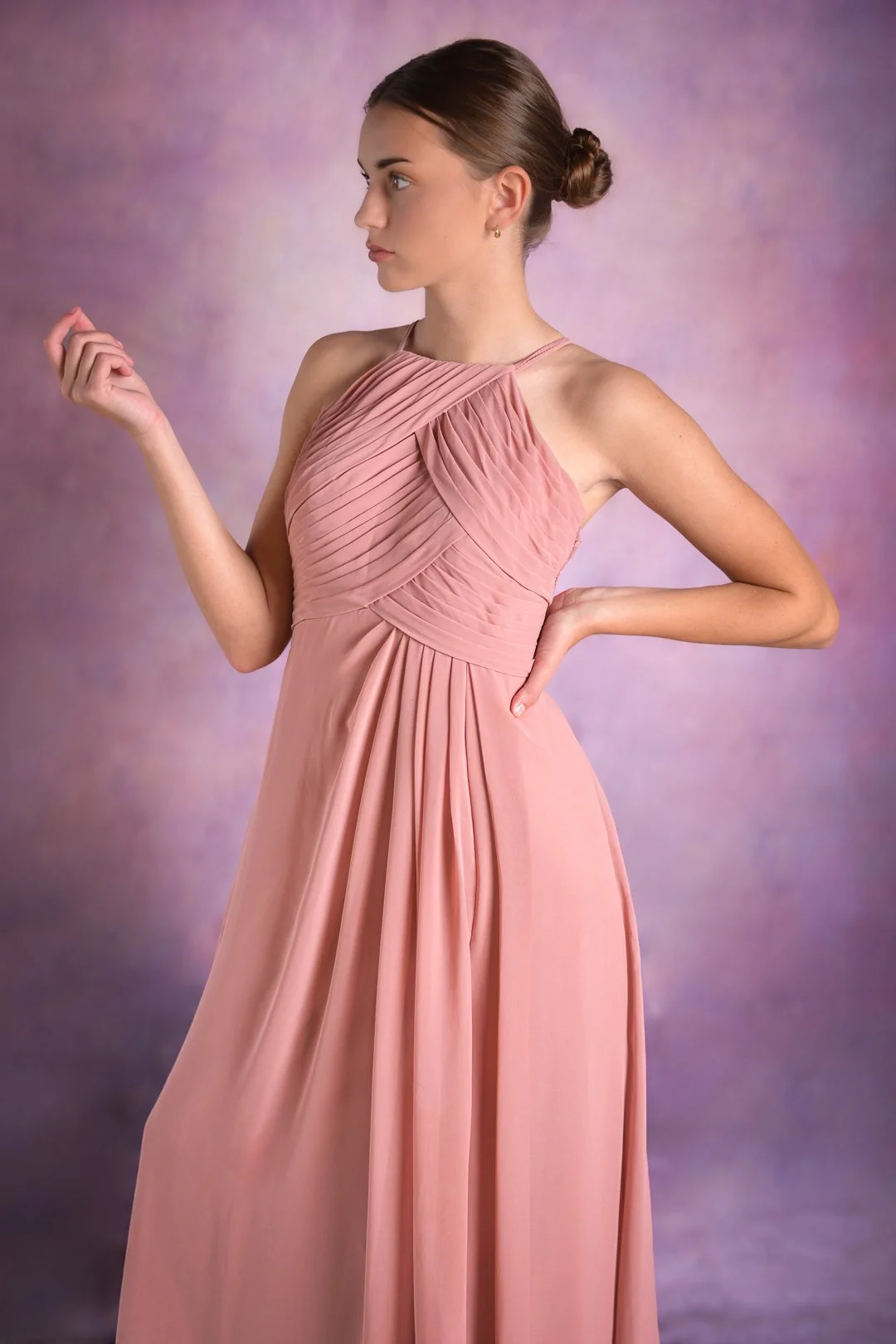 A young woman in a pink, pleated gown with a high neckline and sleeveless design, standing against a soft purple background.