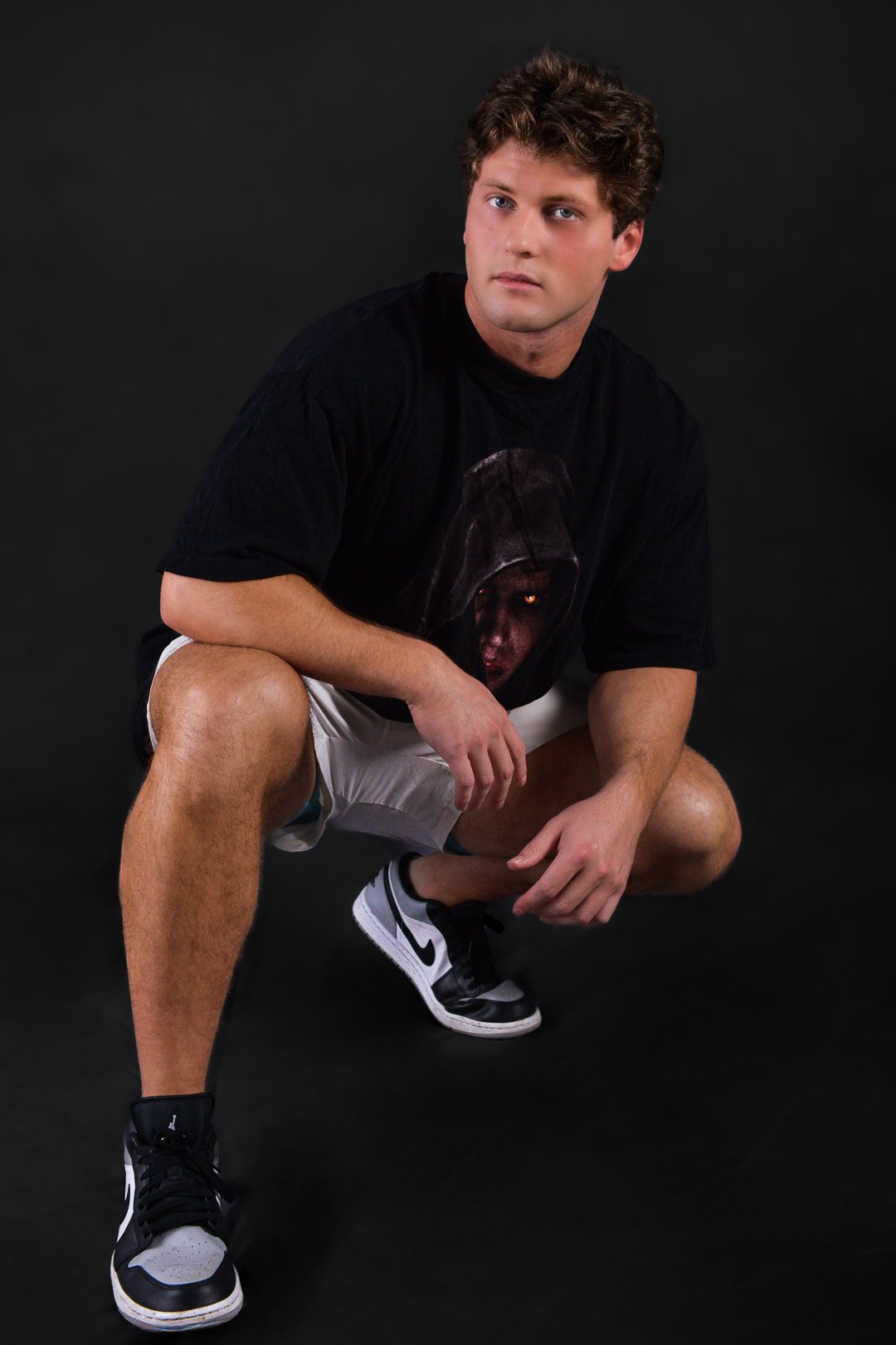 A young man with curly hair squatting on a black background, wearing a black T-shirt with a face in a hooded cloak graphic, beige shorts, and black and white Nike sneakers.