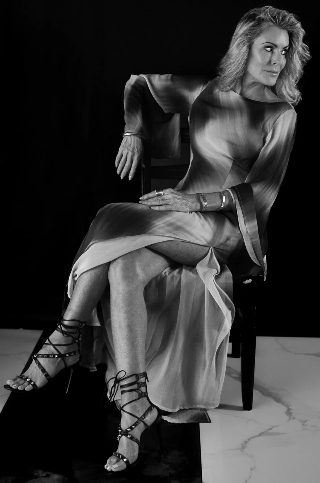 Black and white photo of a woman with wavy blonde hair sitting on a chair, wearing a silky dress and strappy high-heeled shoes, posing with a slightly turned gaze to the side.
