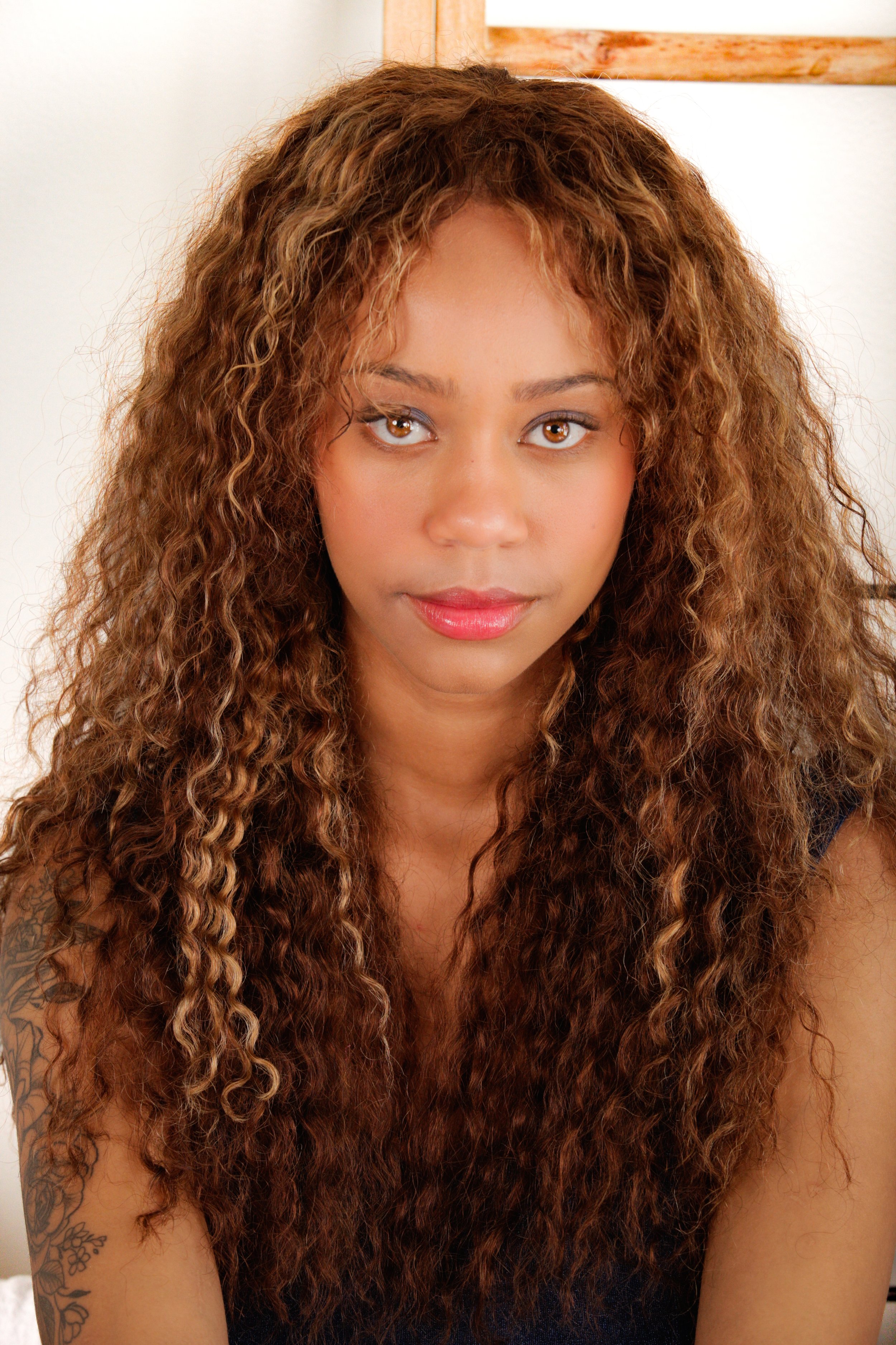 Close-up of a woman with long, curly, reddish-brown hair, light brown skin, and a tattoo on her arm, looking directly at the camera.