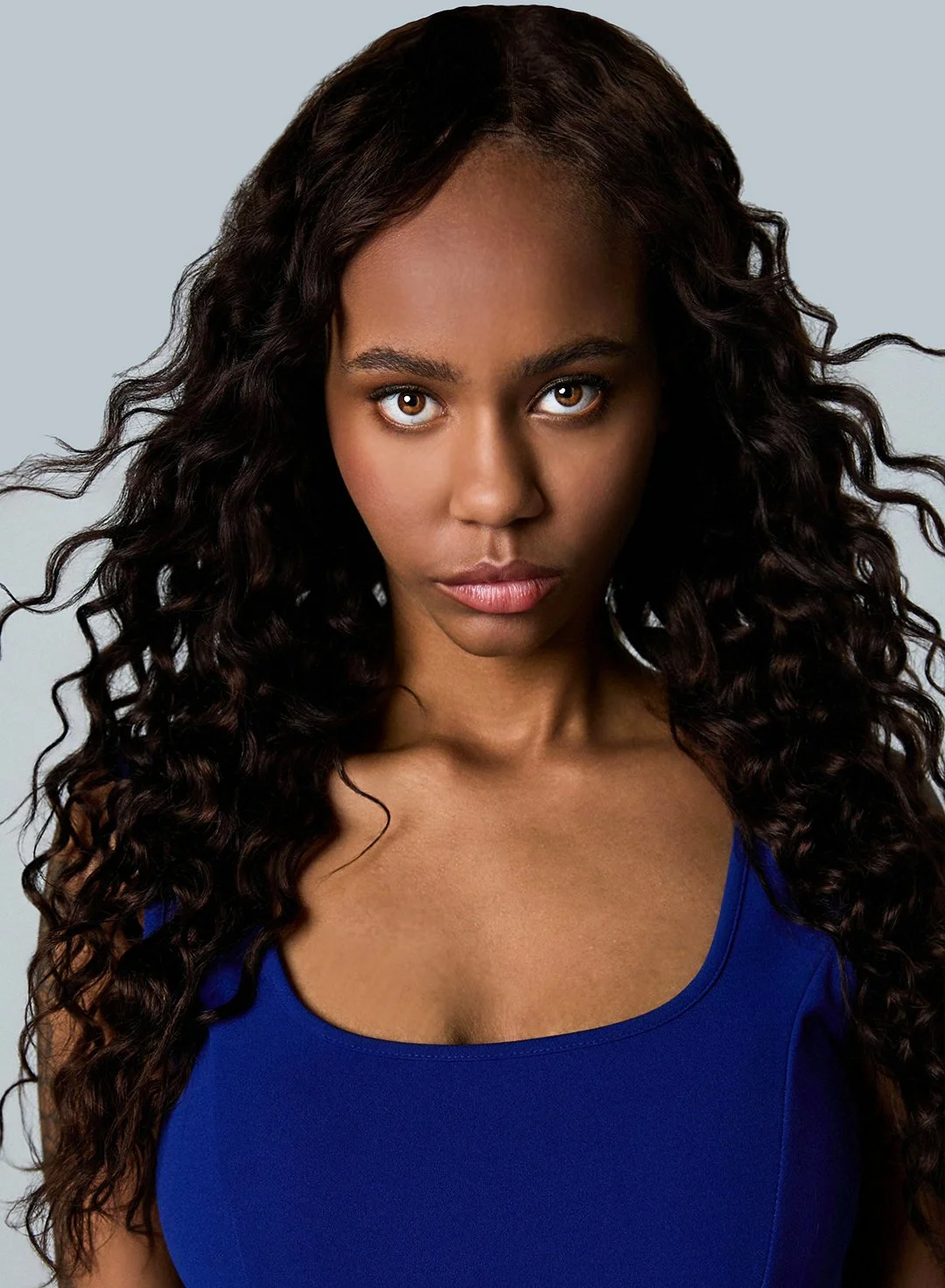 A young woman with dark, curly hair and brown eyes, wearing a blue sleeveless top, looking directly at the camera against a plain gray background.