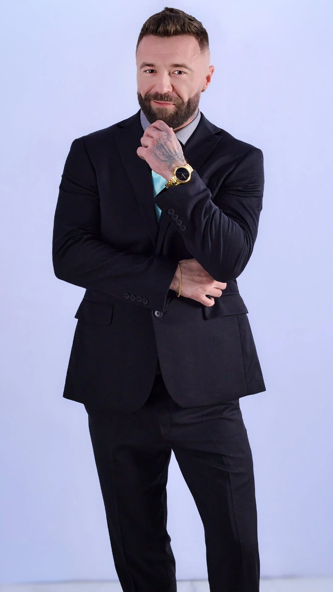 A man in a black suit with a gray shirt and teal tie, sporting a beard and tattooed hand, poses with one hand resting on his chin in front of a plain light blue background.