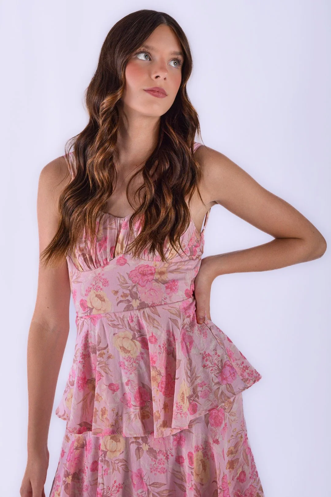 A young woman with wavy brown hair and fair skin wearing a pink floral dress posing with her hand on her hip against a white background.
