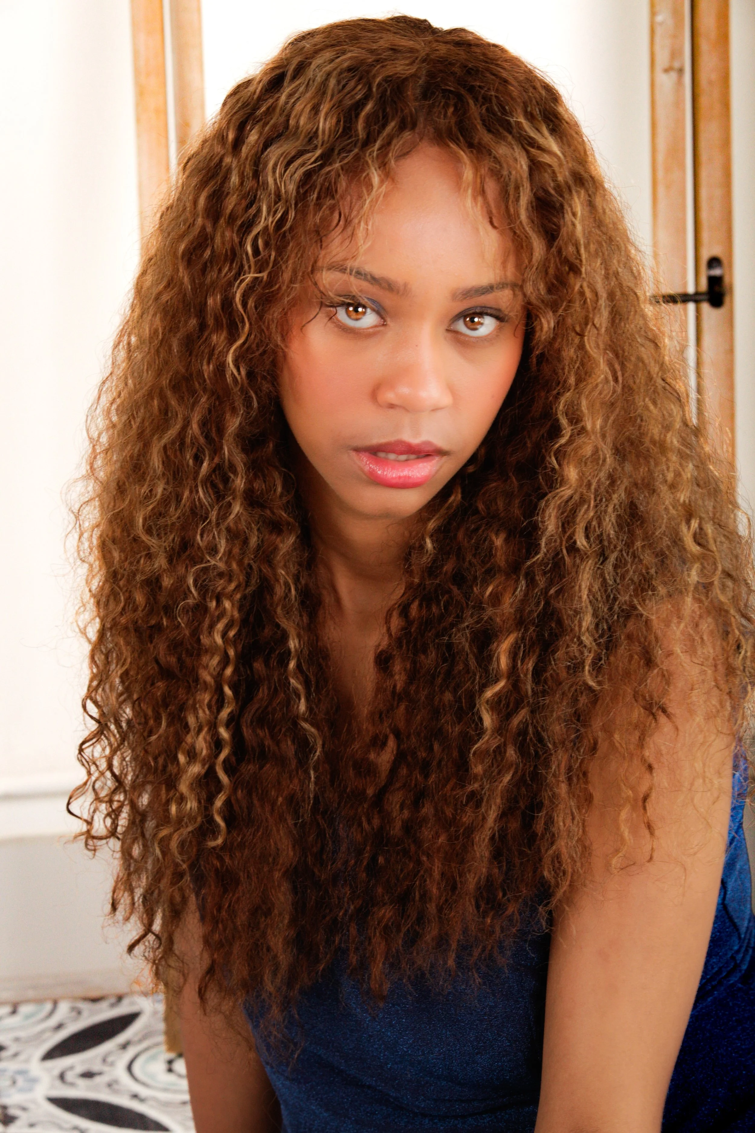 A woman with long, curly brown hair wearing a blue top, looking into the camera with a serious expression.