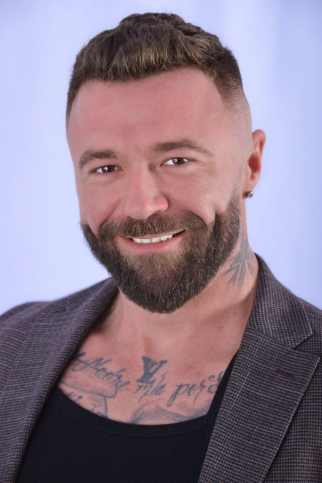 Close-up of a smiling man with a beard, short brown hair, earrings, tattoos on chest, wearing a black shirt and plaid blazer against a light background.