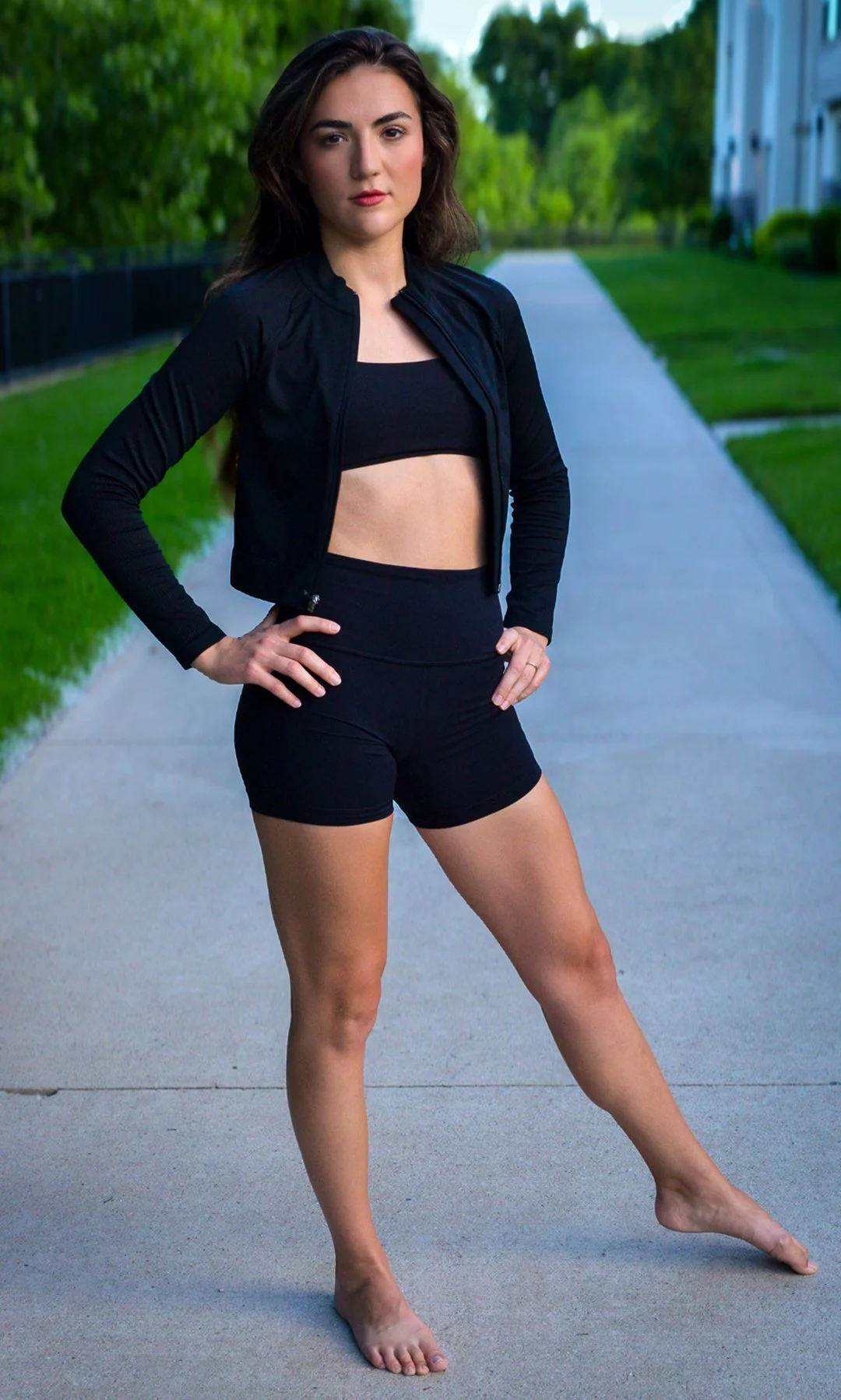A woman standing outdoors on a sidewalk with green trees and a white building in the background, wearing a black sports bra, black shorts, and a black zip-up jacket, with her hands on her hips.