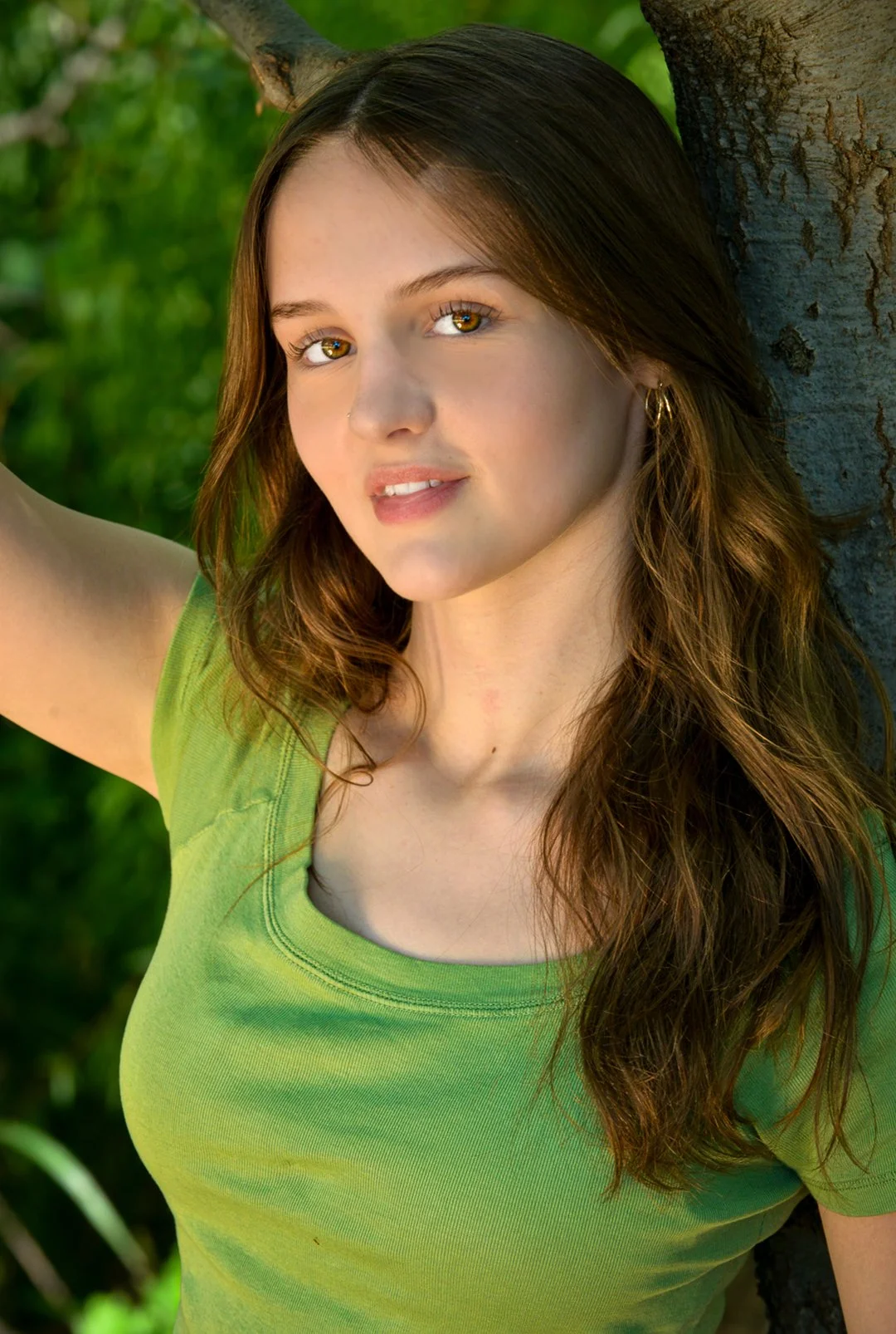 A young woman with long brown hair and hazel eyes leaning against a tree, wearing a bright green t-shirt, in a lush outdoor setting.